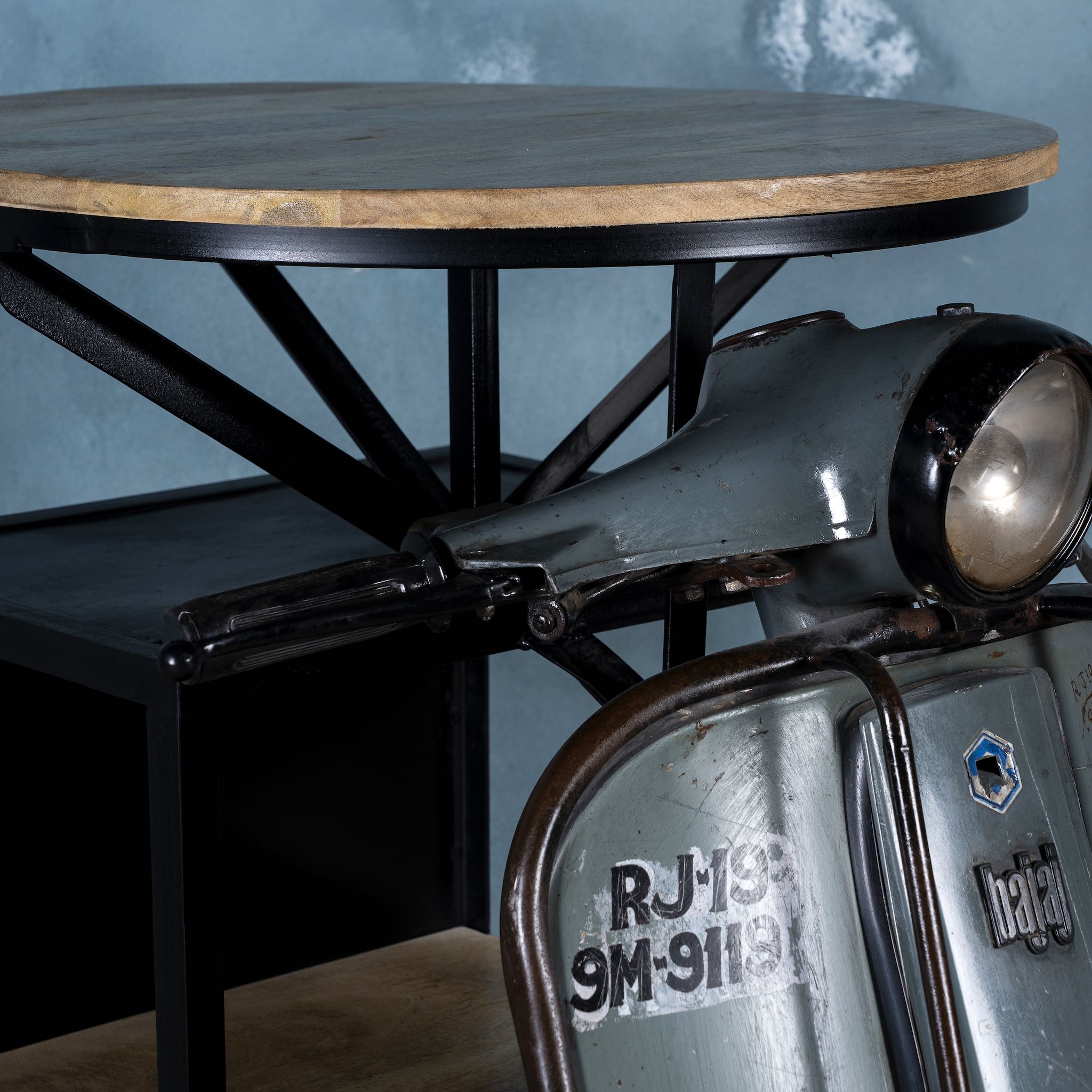 Close up image of a scooter bar table crafted from the front half of an original Bajaj scooter, featuring a round wooden top, metal frame, and rustic industrial finish.