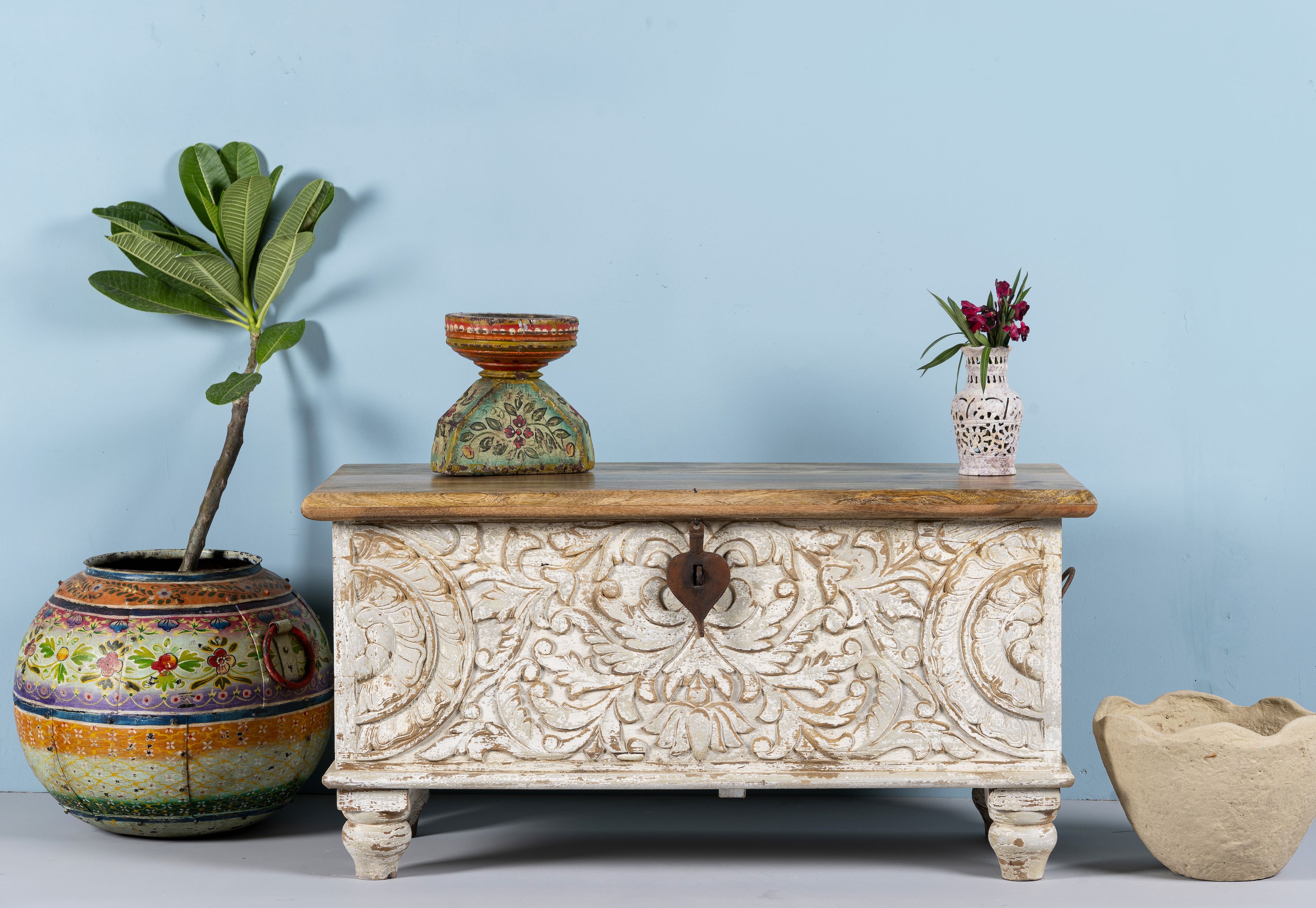 A hand-carved mango wood storage box with floral patterns, a whitewashed finish, and iron handles, displayed against a blue wall with vase and flower arrangement.