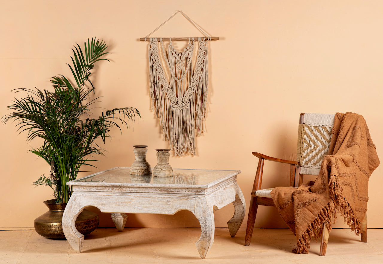 A hand-carved mango wood coffee table with a white finish, accompanied by a glass top, placed in a room with a chair, a potted plant, and a wall hanging decor.