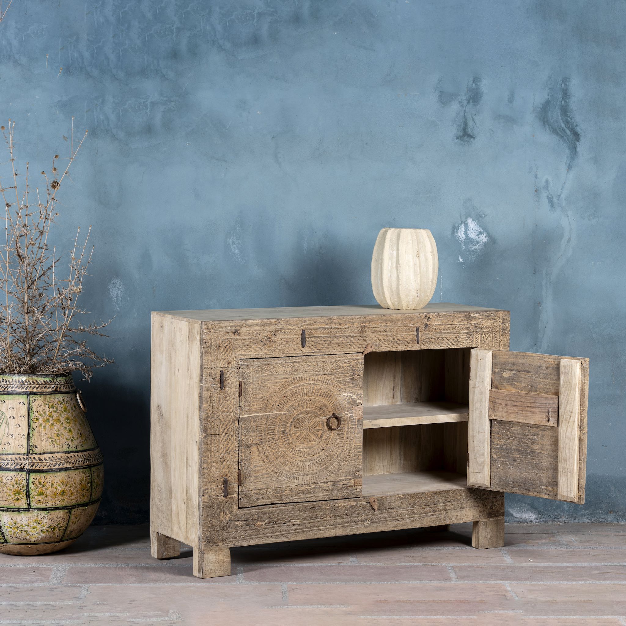 Side angle with open door picture of a sideboard with hand-carved teak wood front, sandblasted for a sun-worn finish — a unique piece in rustic Ibiza style with circular mandala carvings and iron ring handles.