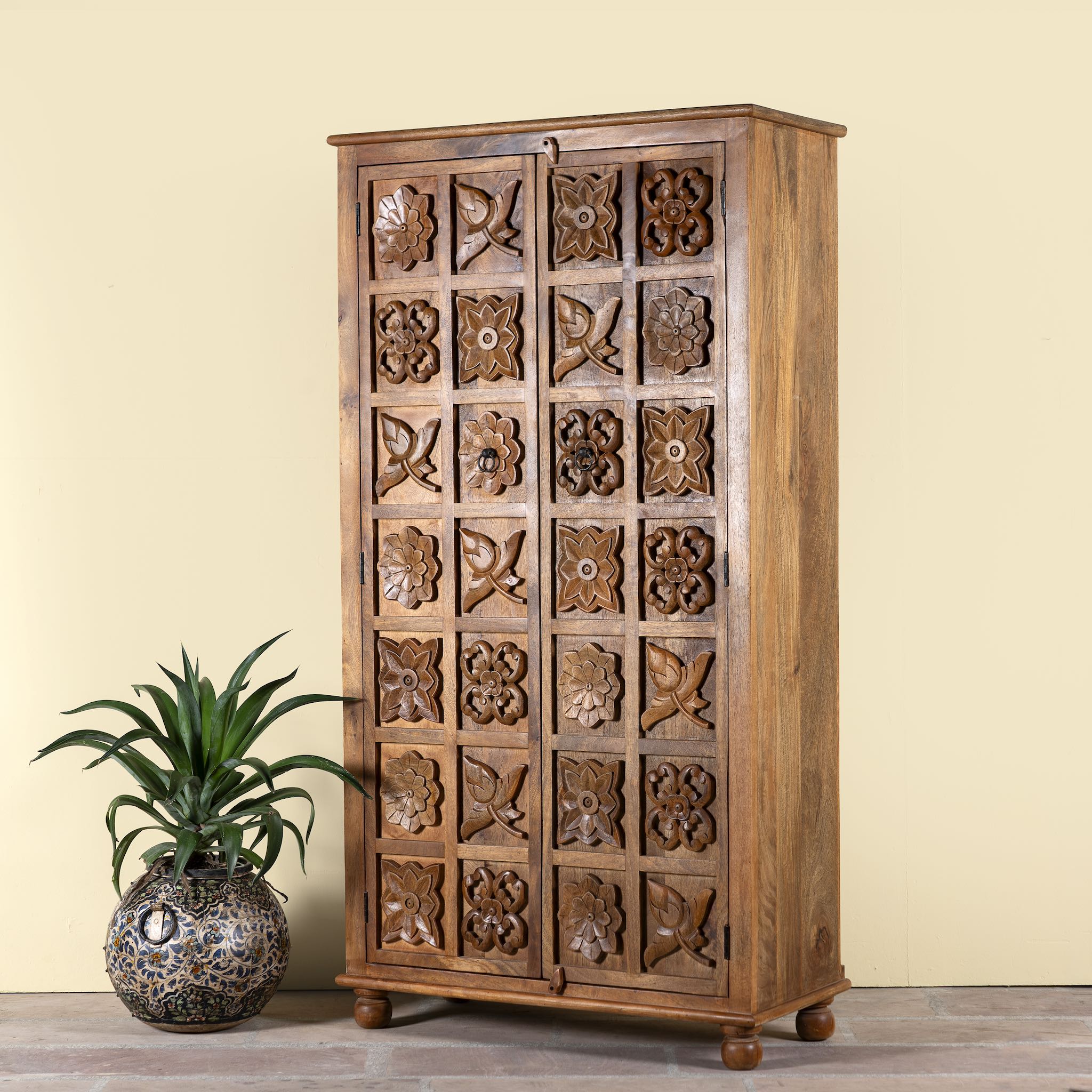Side angle image of a hand-carved mango wood wardrobe with floral motifs; wooden wardrobe in natural finish, 100 × 45 × 190 cm.
