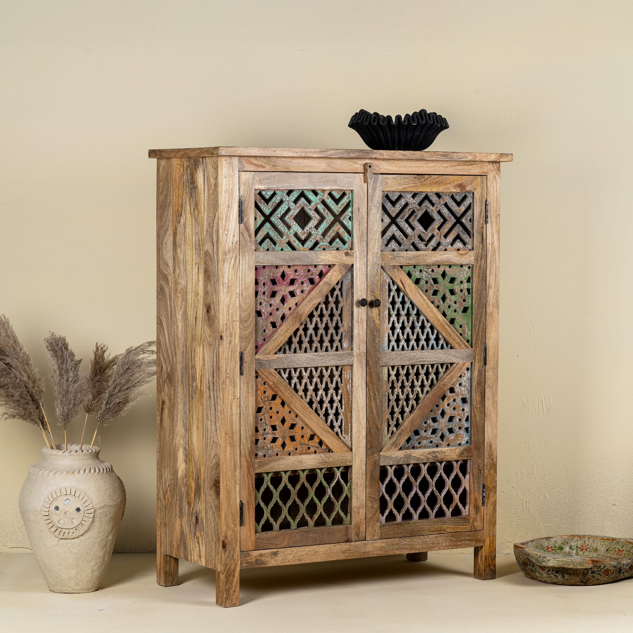 Side angle image of a handmade mango wood cabinet with carved floral and geometric lattice doors, distressed colour accents; artisanal furniture perfect for boho style interiors, 100 × 45 × 130 cm.