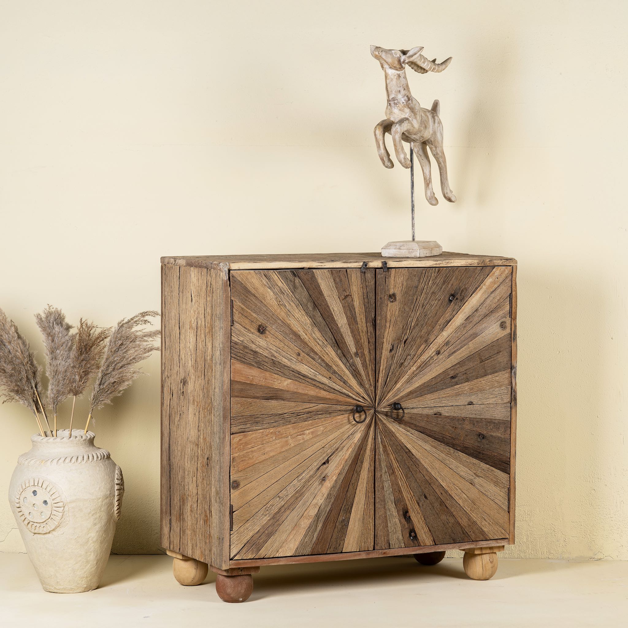 Side angle image of a artistic wooden cabinet in rustic style, made from reclaimed wood with a striking sunburst pattern on the double doors, featuring subtle color variations and round metal ring pulls, elevated on rounded wooden feet.