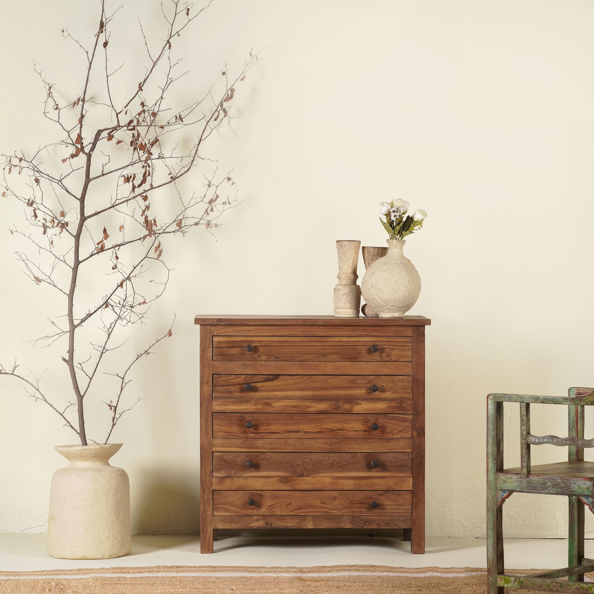 A handcrafted chest of drawers made from recycled teak wood, featuring six drawers with black metal knobs, positioned against a neutral background with decorative items on top.