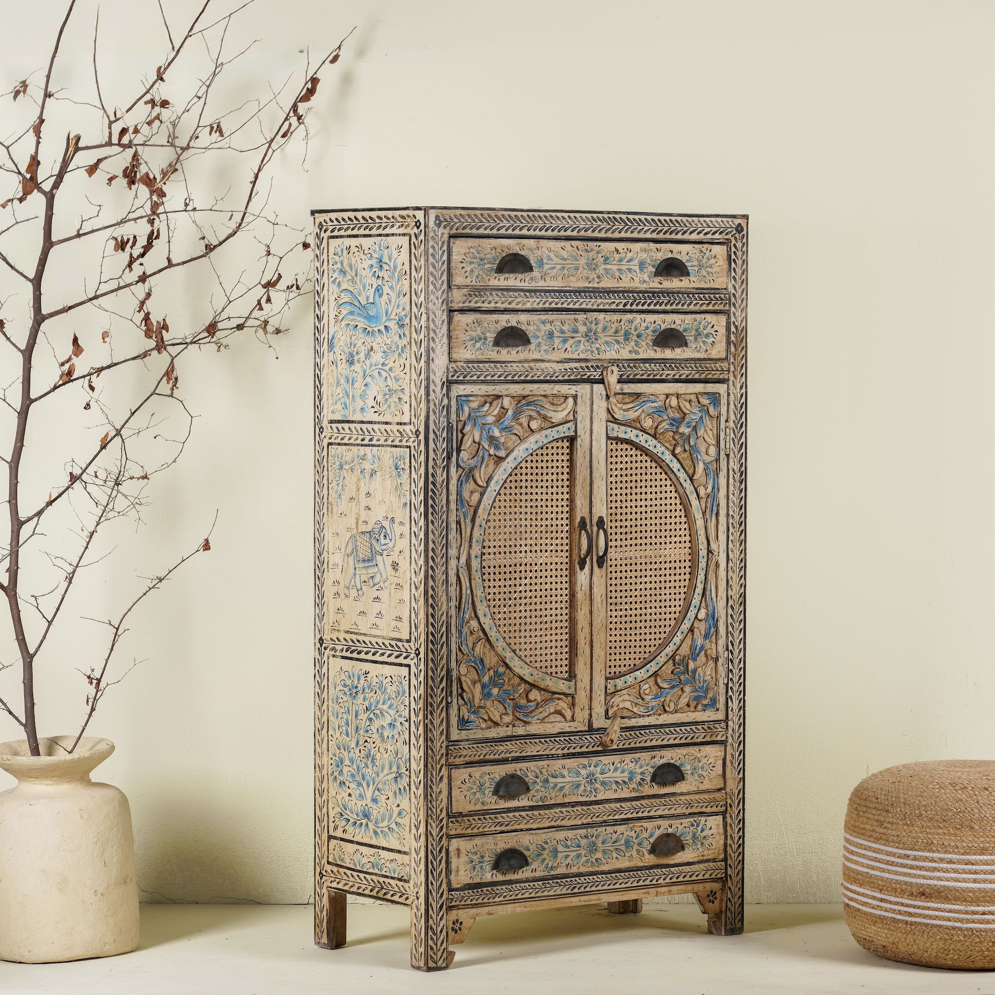 Side angle image of a hand-painted cabinet made from mango wood, featuring rattan inlay doors framed by a carved circular motif, floral blue and green accents, and five drawers for additional storage.
