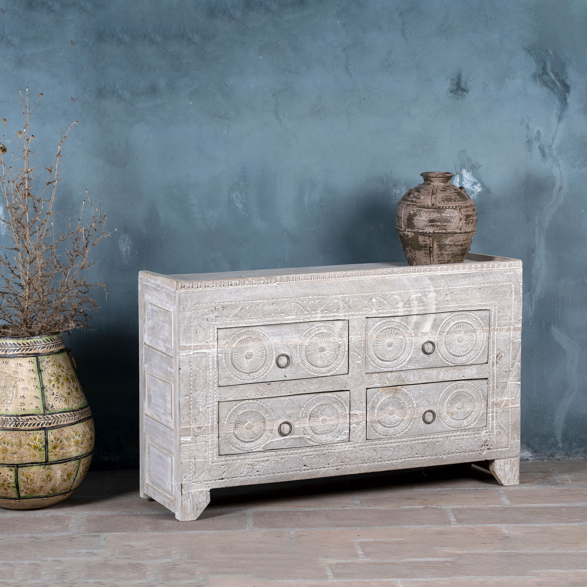 Side angle picture of a Ibiza style sideboard with six drawers, made from hand-carved wood in a whitewashed finish, featuring circular mandala motifs and rustic iron handles.