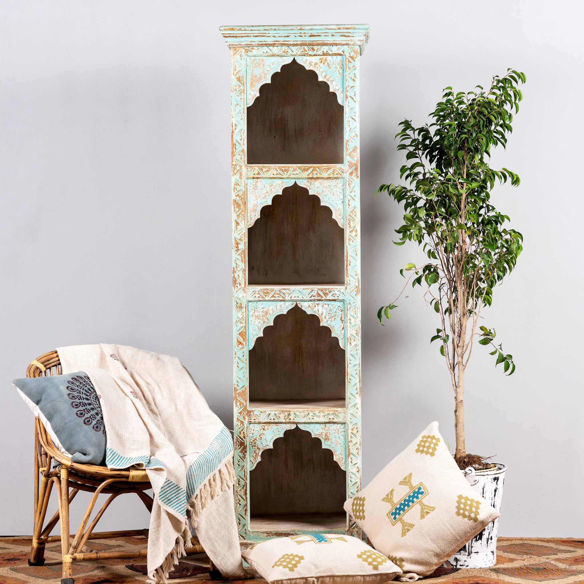 A tall hand-carved mango wood shelf with distressed pastel blue finish, featuring four open shelves, placed next to a chair with a plant and cushions.