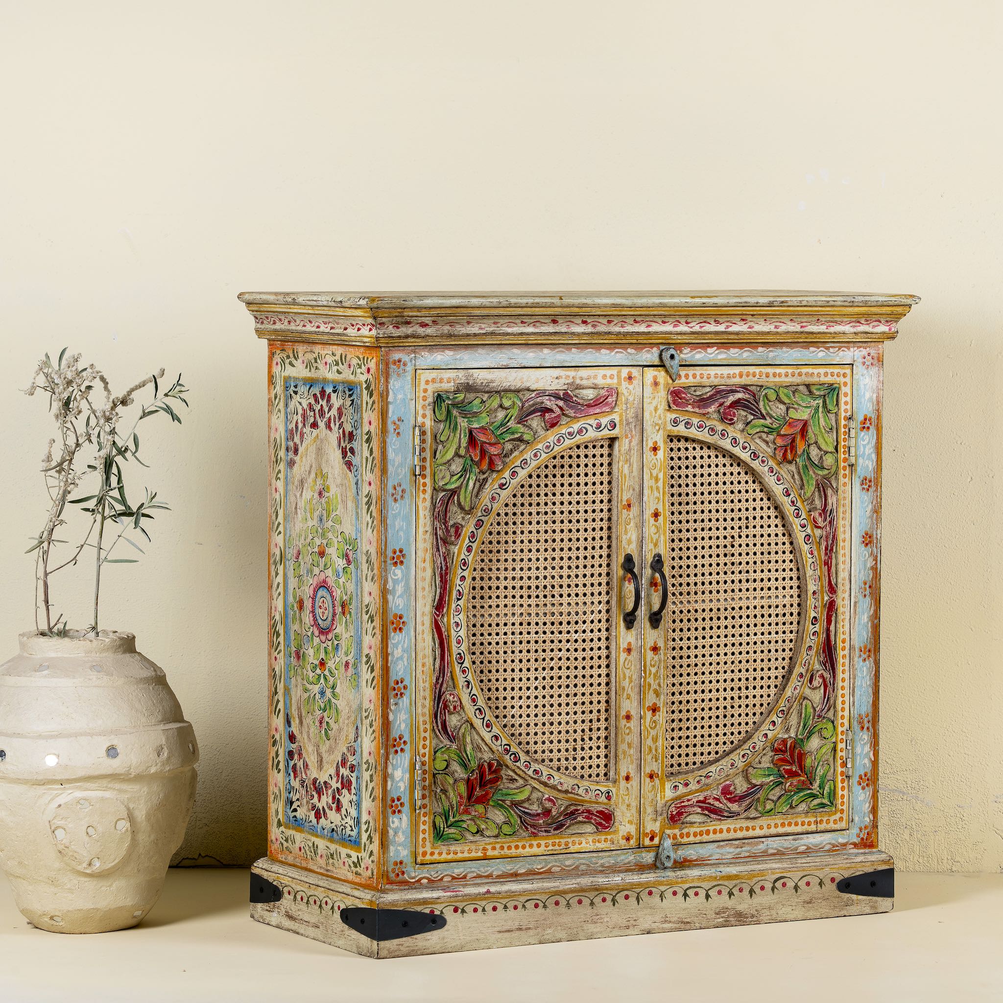 Side view of a hand-painted, hand-carved cupboard made of mango wood with floral design and rattan inserts.