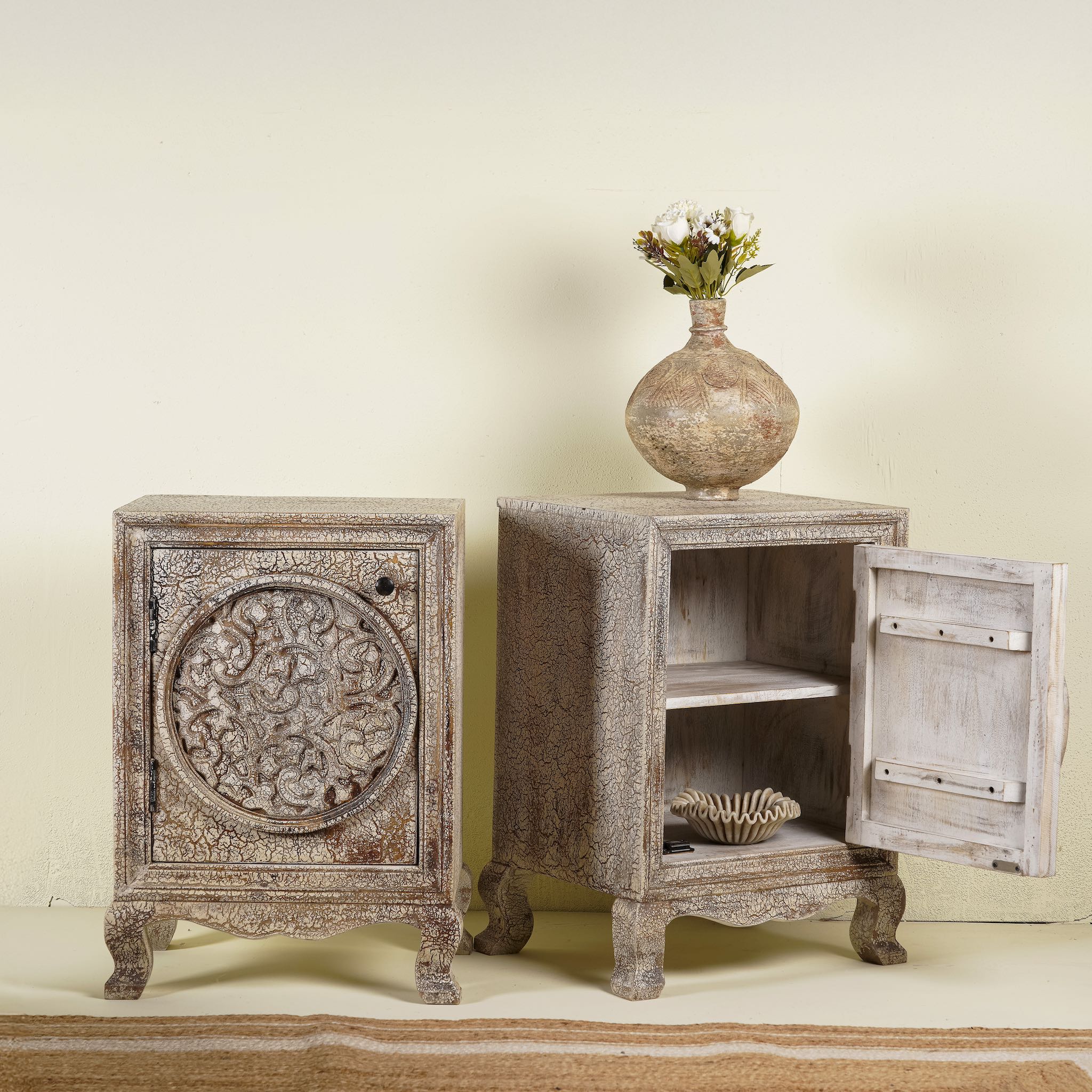 Open door picture of a pair of hand-carved bedside tables made of mango wood, featuring intricate round floral motifs on the doors, crackled vintage finish, and elegant curved legs in a distressed antique design.