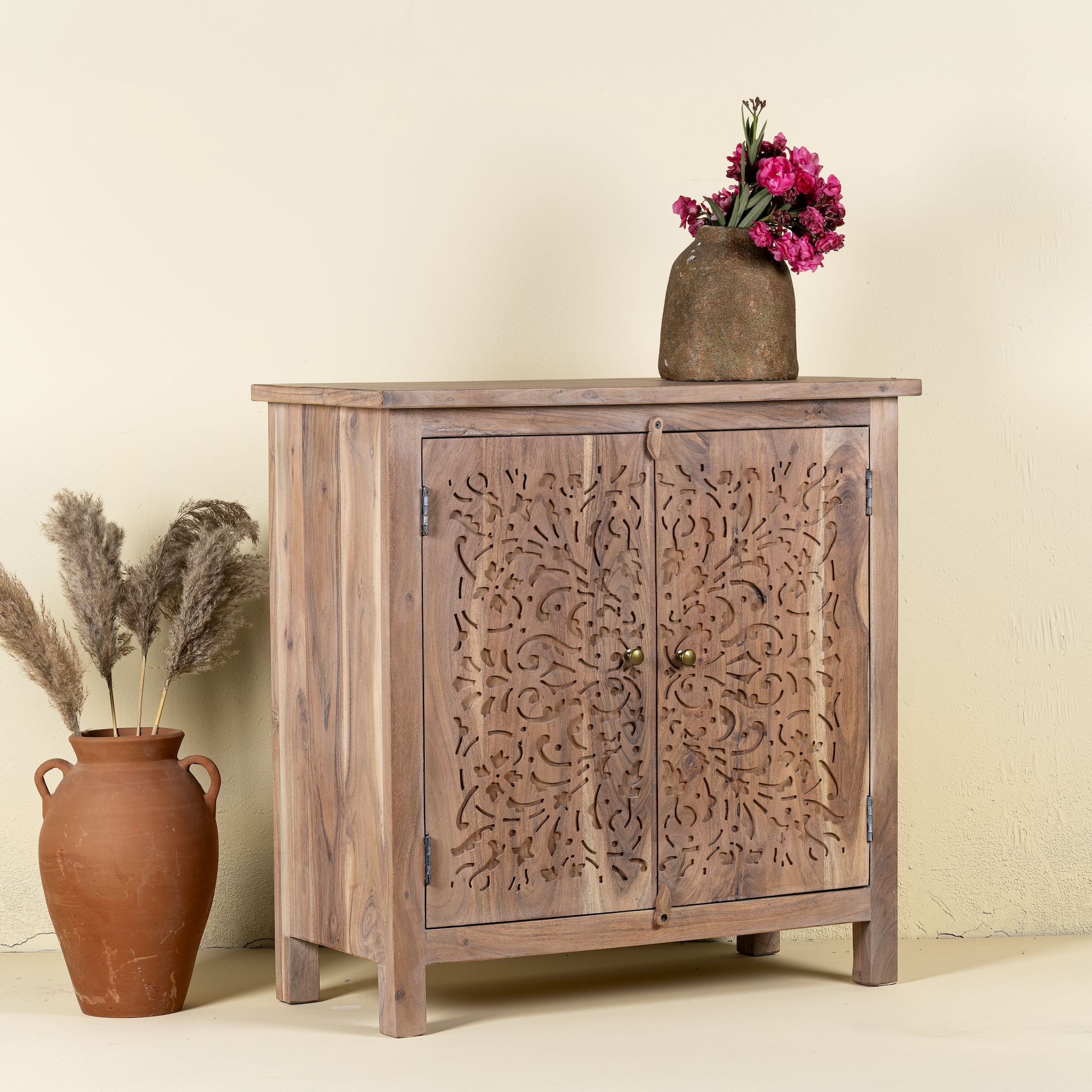 Side angle image of a carved acacia wood cupboard with floral cutout doors and brass knobs, 90 × 40 × 90 cm.