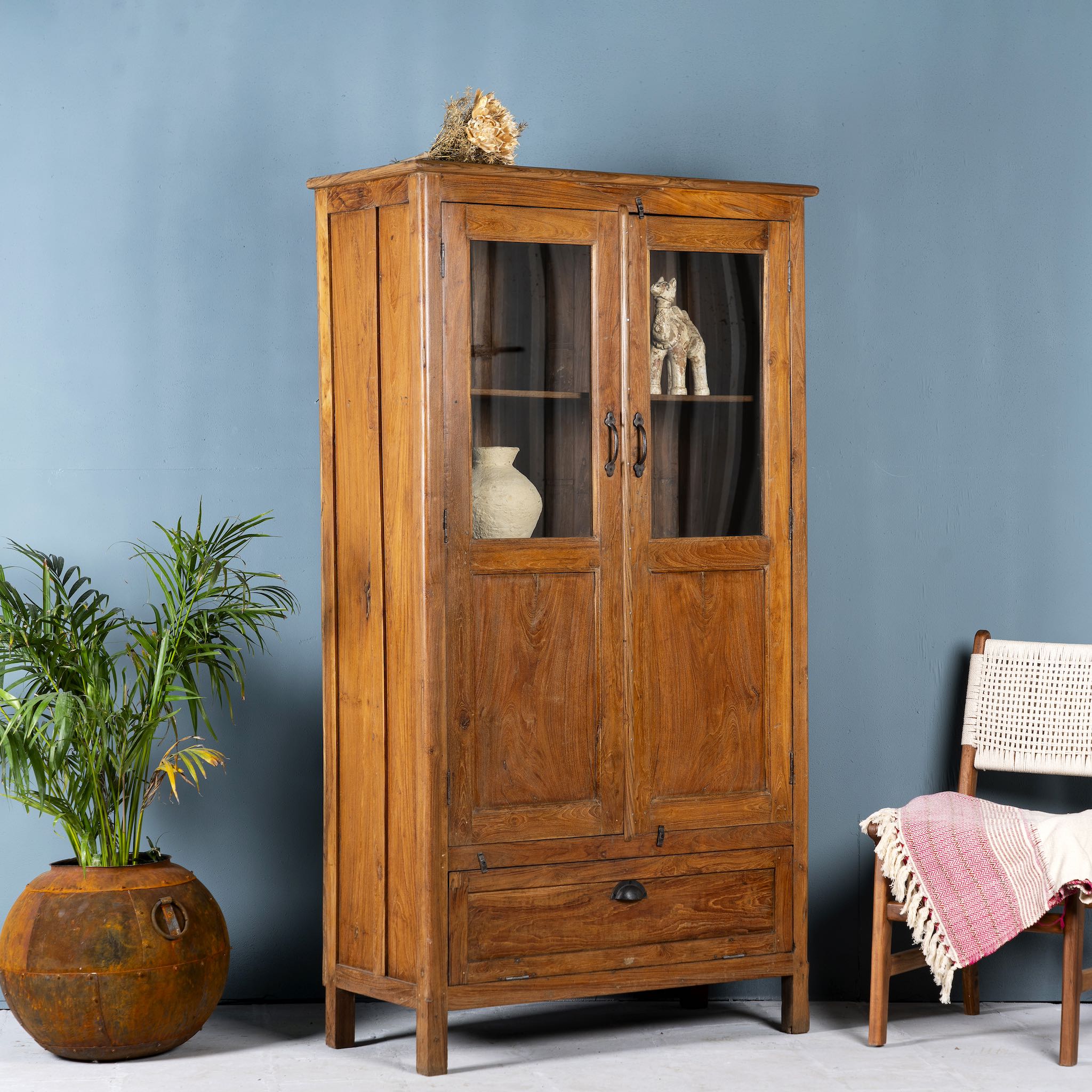Side angle image of a tall reclaimed teak wood display cabinet with two glass doors and a bottom drawer – wooden display cabinet crafted as sustainable furniture with natural elegance.