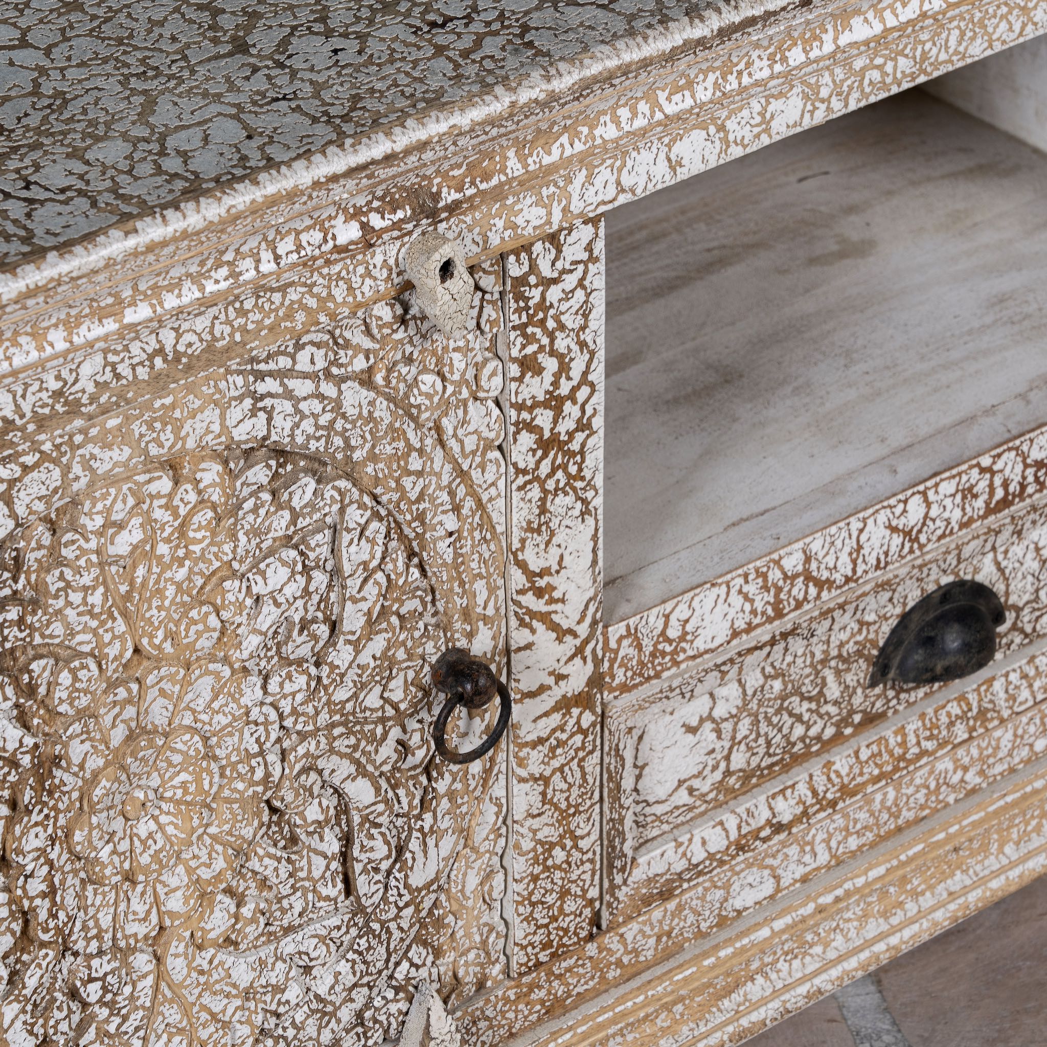 Close up image of a carved mango wood lowboard with floral doors, crackled vintage finish, drawer, and open shelf, 150 × 40 × 64 cm.