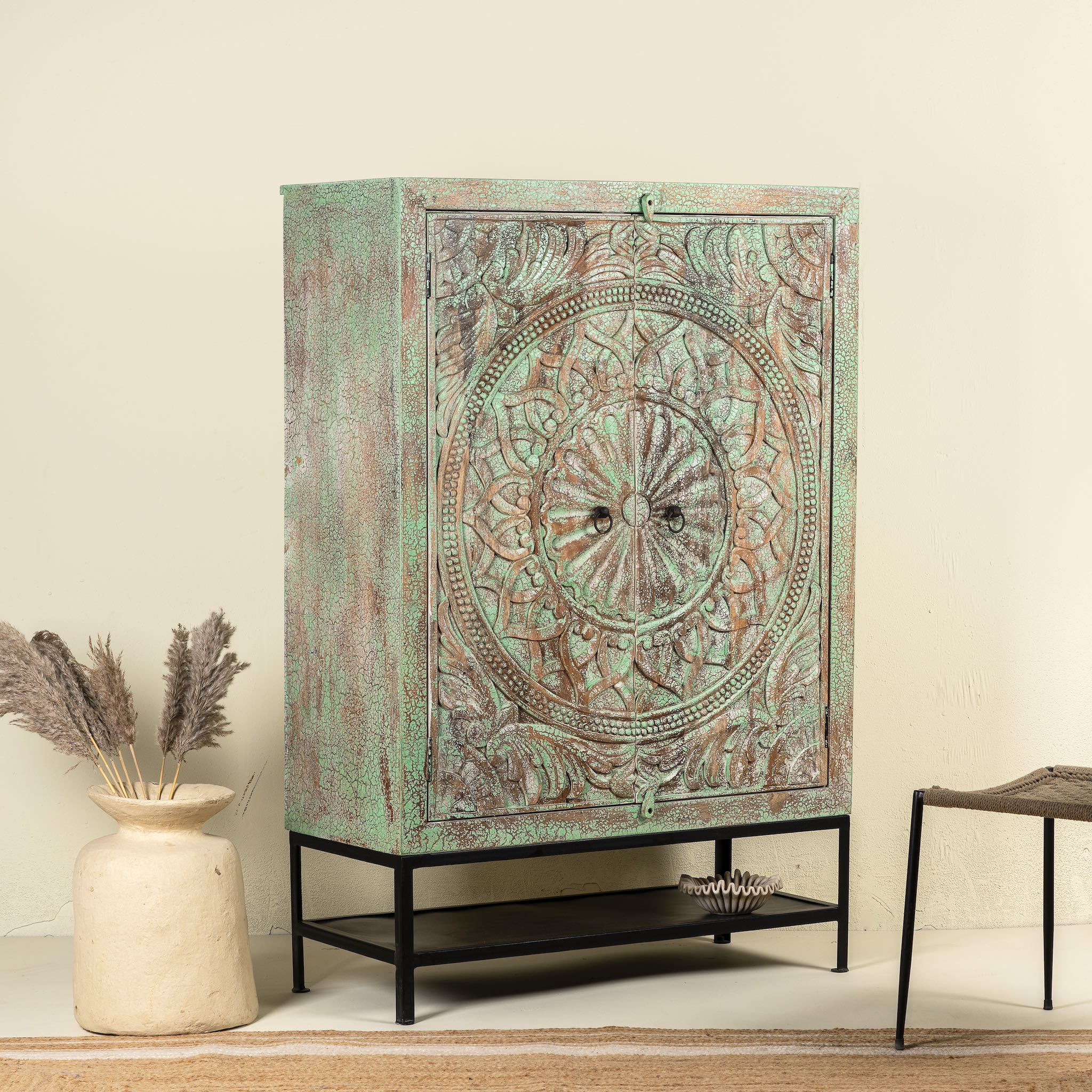Side angle image of a carved mango wood cabinet with floral hand-carving, distressed green finish, iron stand, 100 × 40 × 150 cm.
