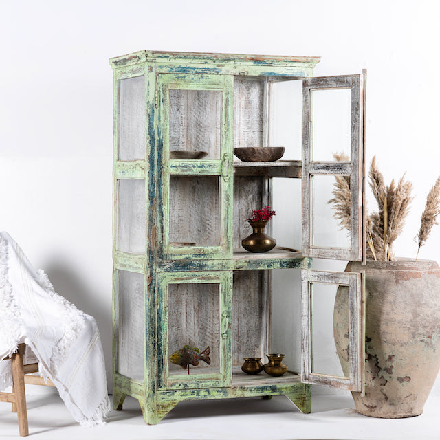 Side angle image with open door of a refurbished teak wood showcase with distressed green finish – sustainable furniture with boho style interior appeal and glass-panelled storage.