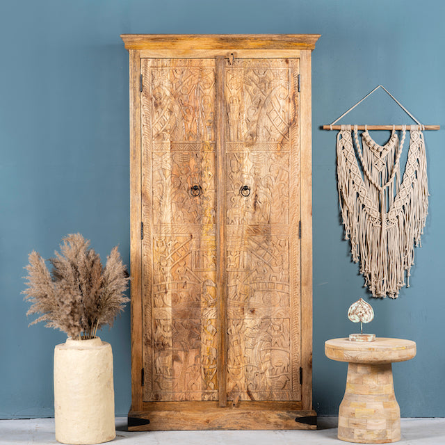 A hand-carved mango wood cabinet with traditional village scenes, Ibiza style, against a blue background, accompanied by a decorative wall hanging, a vase with dried plants, and a decorative object on top of the cabinet.