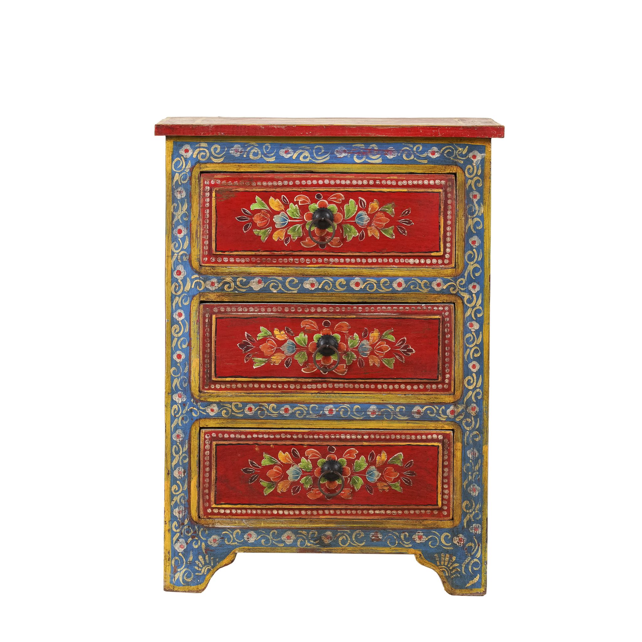 White background image of a hand-painted reclaimed wood nightstand with vibrant floral motifs, featuring three drawers and a recycled wood top, placed next to a vase with dried plants.