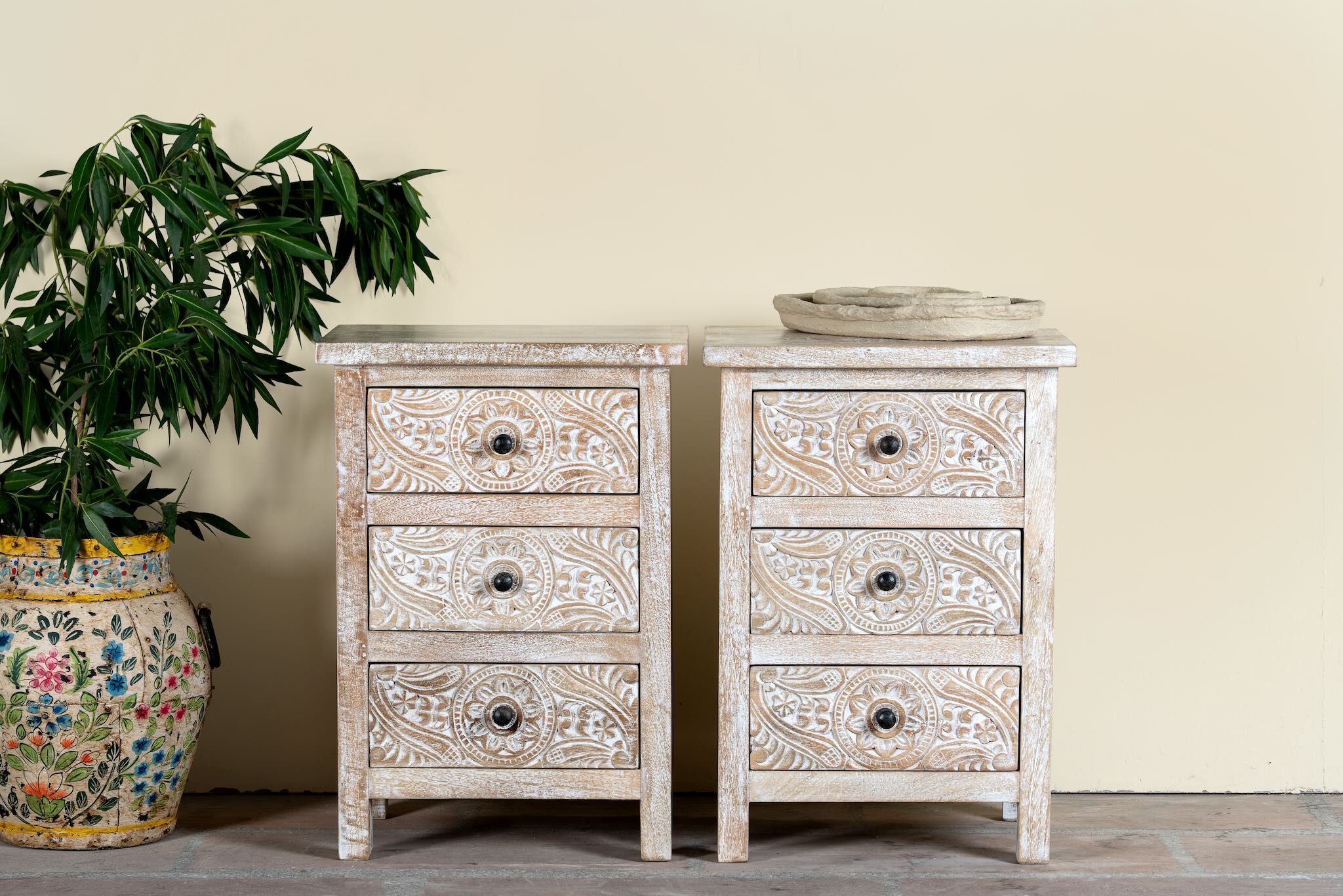 Pair of mango wood nightstands Ved in a whitewashed vintage finish, featuring hand-carved drawer fronts with ornate circular and leafy motifs. Stylish and compact vintage-style bedside tables with three drawers for elegant storage.