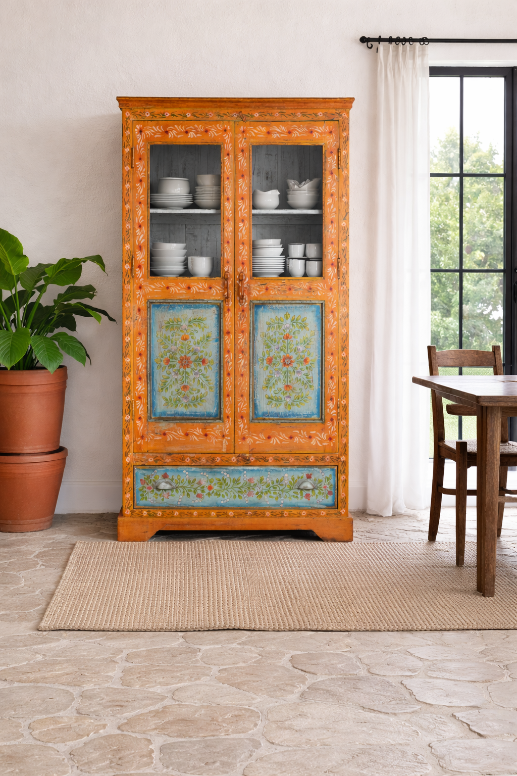 Lifestyle imagination of a of a hand-painted teak wood showcase with floral motifs and glass doors – sustainable furniture crafted from recycled wood with traditional Indian artistry.
