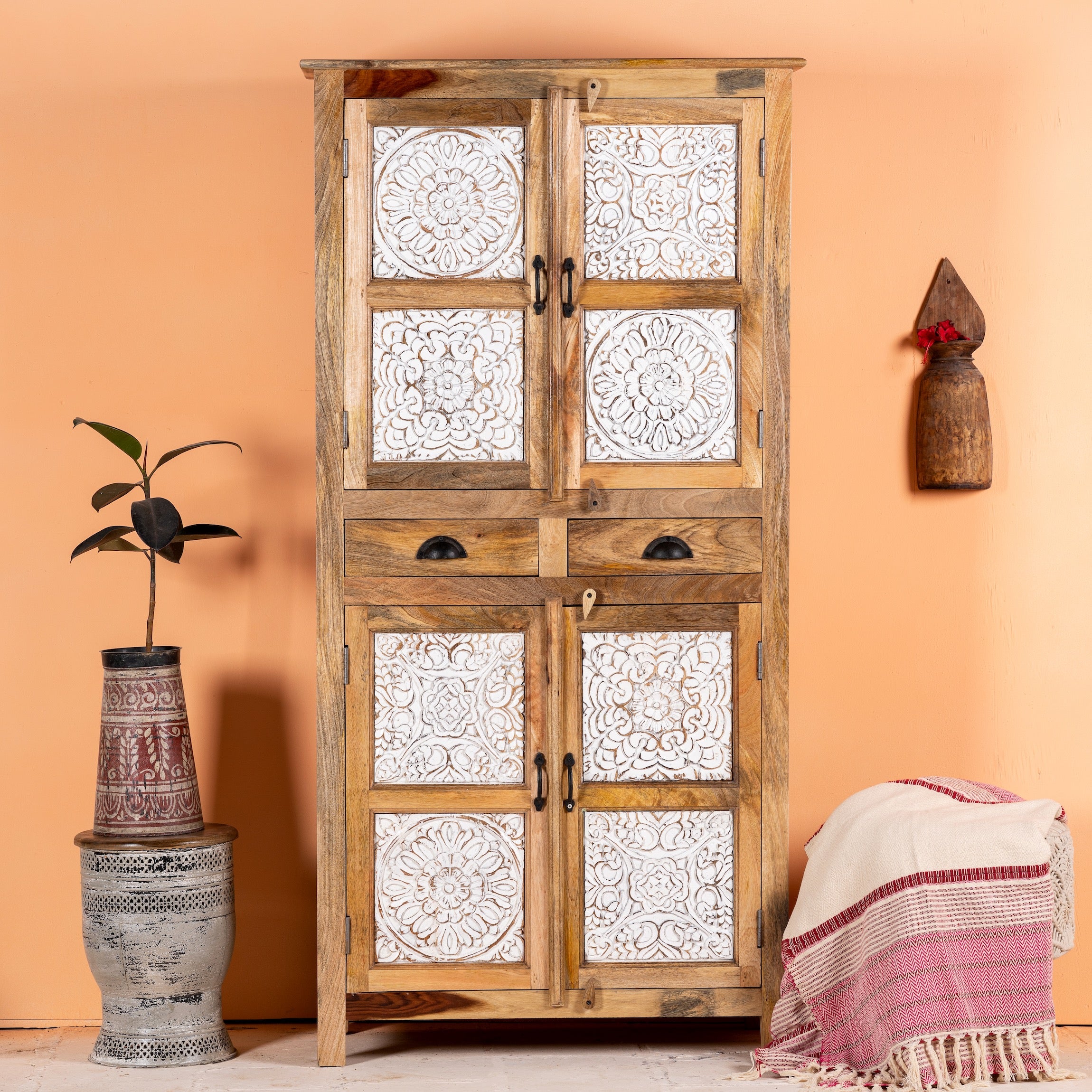 Hand-carved mango wood wardrobe with whitewashed floral patterns and natural wood finish, featuring two drawers and four compartments, displayed in a home setting with a potted plant and a traditional textile adorned the foreground.