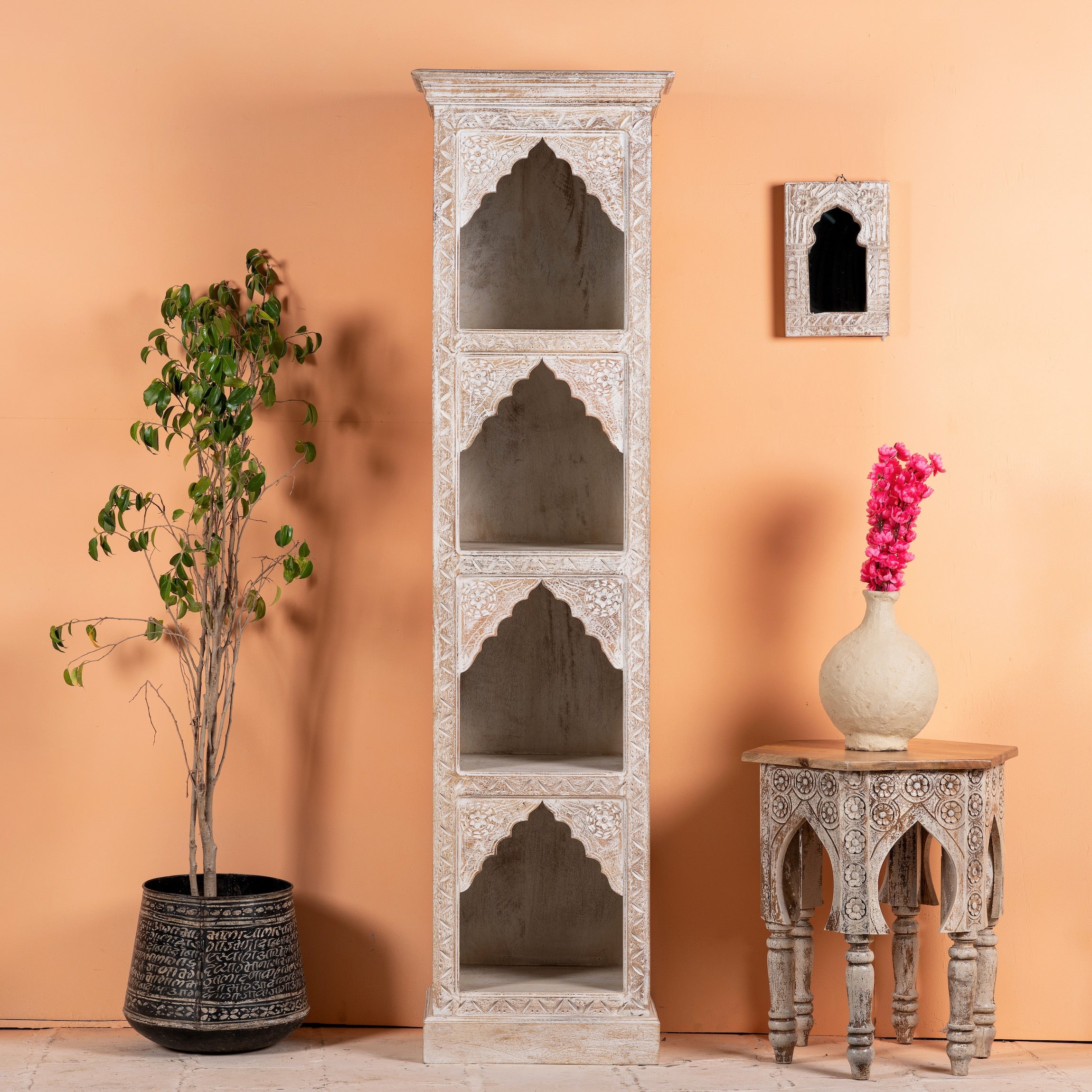 A white, hand-carved mango wood Hooghly bookshelf with asymmetrical designs, placed against a pink wall with a decorative object on top and a potted plant to the side.