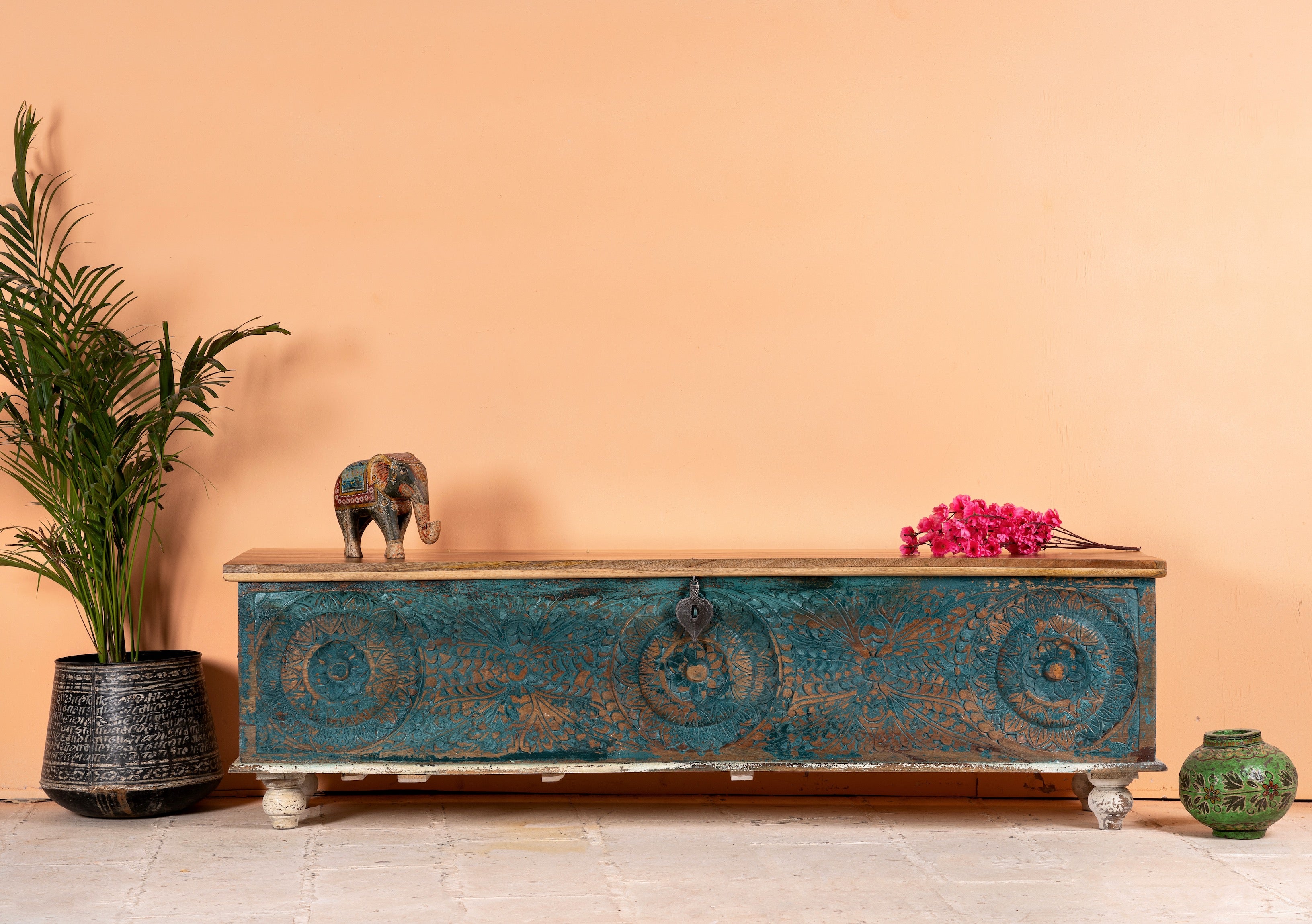 A blue and brown wooden storage chest with intricate hand-carved patterns, placed against an orange wall with decorative items on and around it.