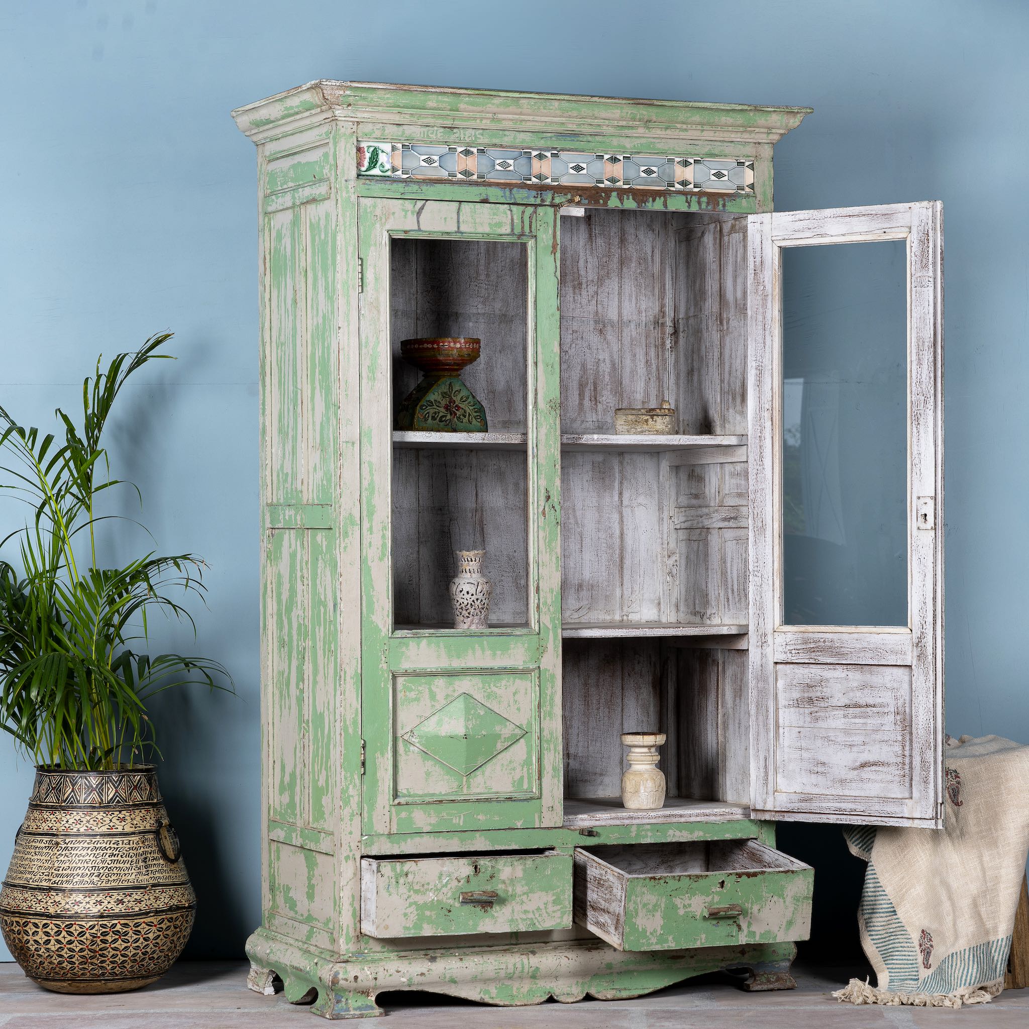 Side angle image with open door of a reclaimed teak wood vitrine with distressed green finish, ceramic inlays, and glass-panelled doors – sustainable furniture with vintage Indian charm.