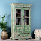 Side angle image of a reclaimed teak wood vitrine with distressed green finish, ceramic inlays, and glass-panelled doors – sustainable furniture with vintage Indian charm.