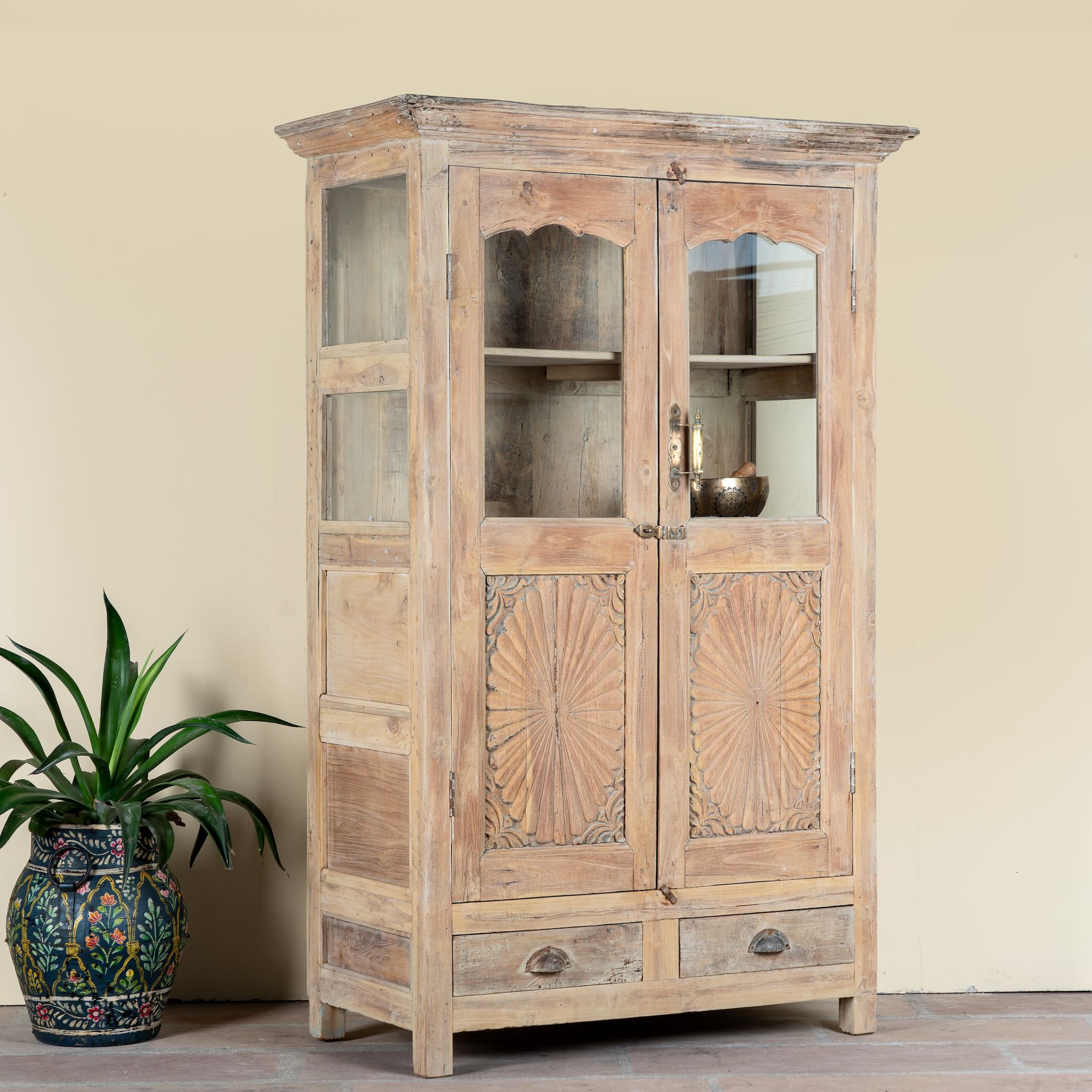 Side angle image of a reclaimed teak wood showcase Aasta with Mediterranean design, featuring hand-carved doors, glass panels, and drawers. A unique piece of refurbished Indian furniture offering elegant display and practical storage.