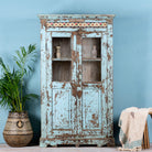 A light blue, antique teak wood showcase with ceramics, featuring two glass doors and multiple shelves, placed against a blue wall with a palm plant and a traditional textile behind it.