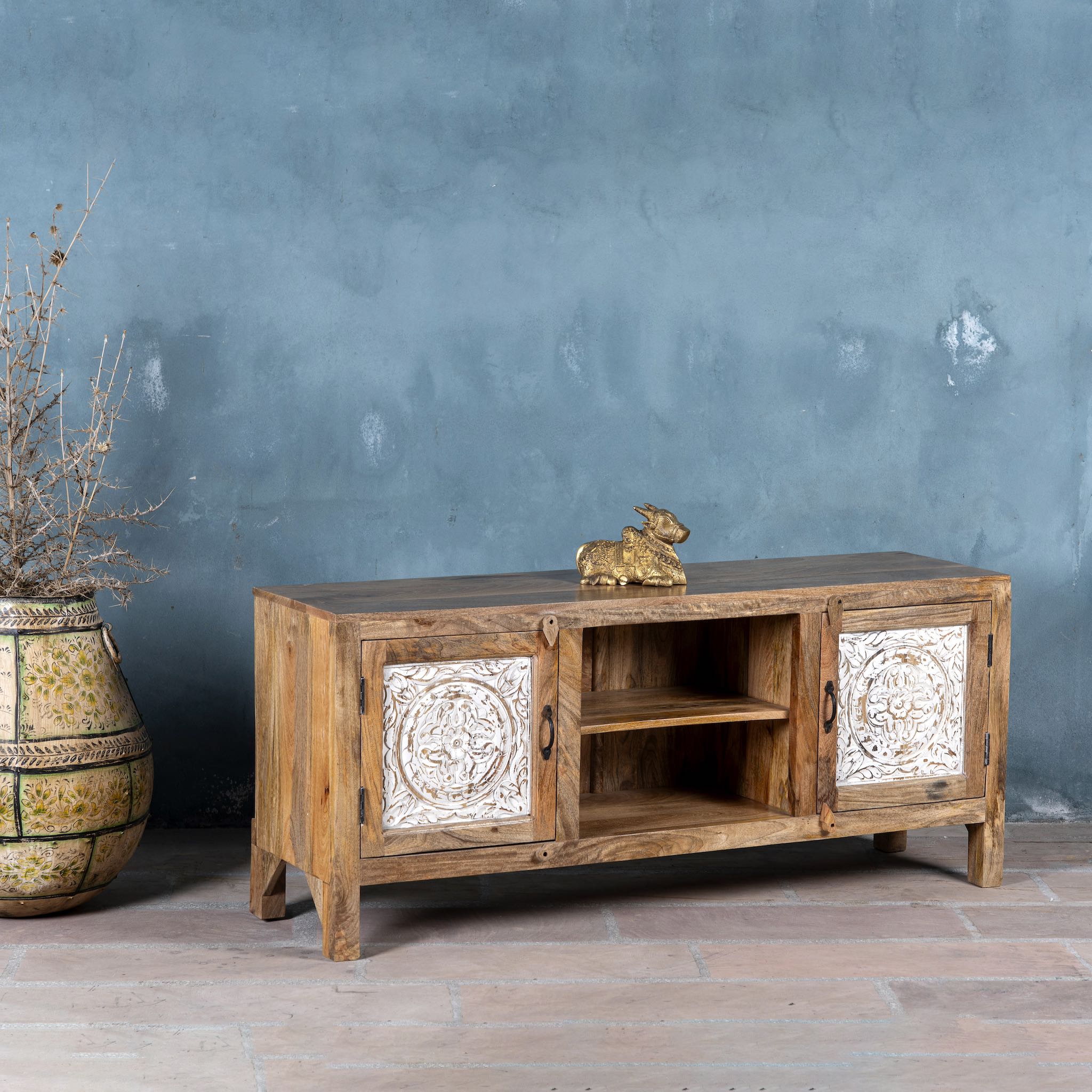 Side angle view of a mango wood low board featuring hand-carved floral motif doors with whitewashed detailing, open central shelving, and a warm natural wood finish.