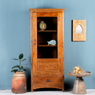 A refurbished antique Indian teak showcase with a glass door and a drawer, featuring interior shelves, placed against a blue wall with decorative objects on the shelves and a plant beside it.