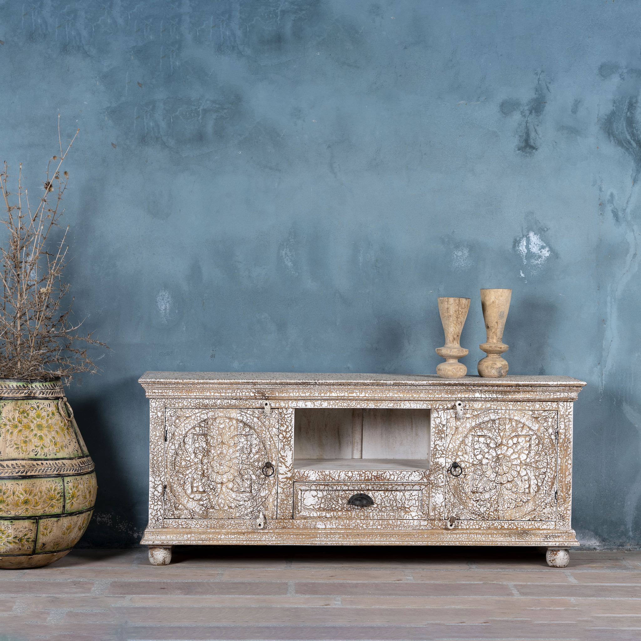 Low board made from solid mango wood with intricately hand-carved circular floral doors, distressed white finish, and central open shelf with drawer for functional storage.