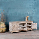 Side angle view of a low board made from solid mango wood with intricately hand-carved circular floral doors, distressed white finish, and central open shelf with drawer for functional storage.