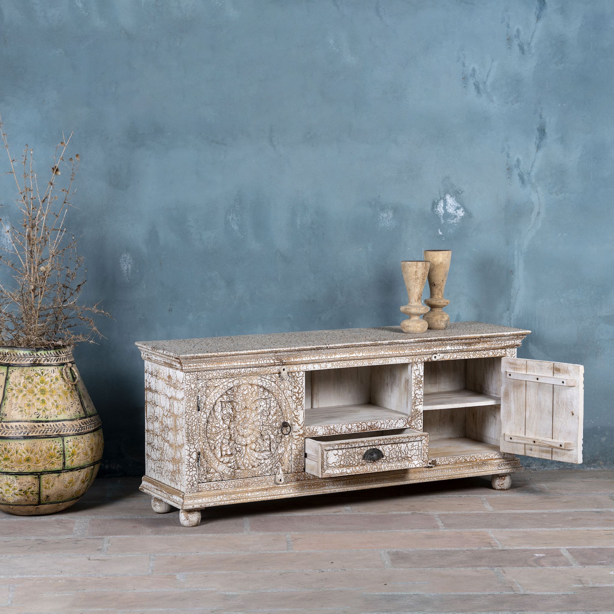 Side angle view of a low board made from solid mango wood with intricately hand-carved circular floral doors, distressed white finish, and central open shelf with drawer for functional storage.