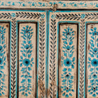 Close up image of a tall cabinet made from reclaimed wood, featuring hand-painted floral motifs in soft blue tones on panelled doors and drawers, with a distressed finish that adds vintage charm and highlights its artisanal craftsmanship.
