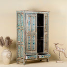 Open door image of a tall cabinet made from reclaimed wood, featuring hand-painted floral motifs in soft blue tones on panelled doors and drawers, with a distressed finish that adds vintage charm and highlights its artisanal craftsmanship.
