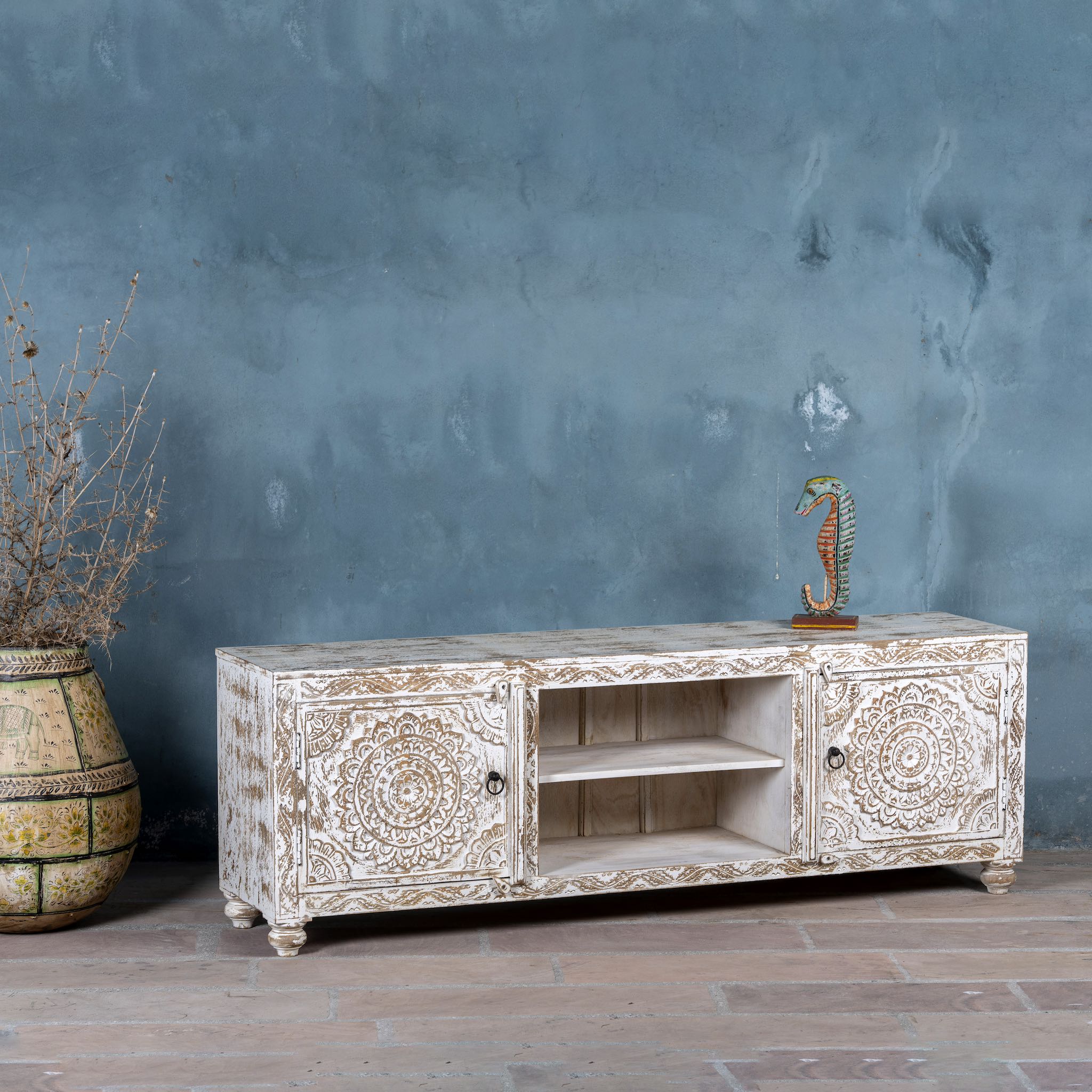 Side angle of a low board made from solid mango wood, featuring intricate hand-carved mandala motifs, whitewashed finish, and practical storage with two doors and open shelving.