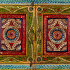 Close up image of a small hand-painted console made from reclaimed wood, featuring vibrant decorative motifs in red, green, blue, and gold on double doors, with intricate floral and geometric patterns and a lower open shelf for added display.
