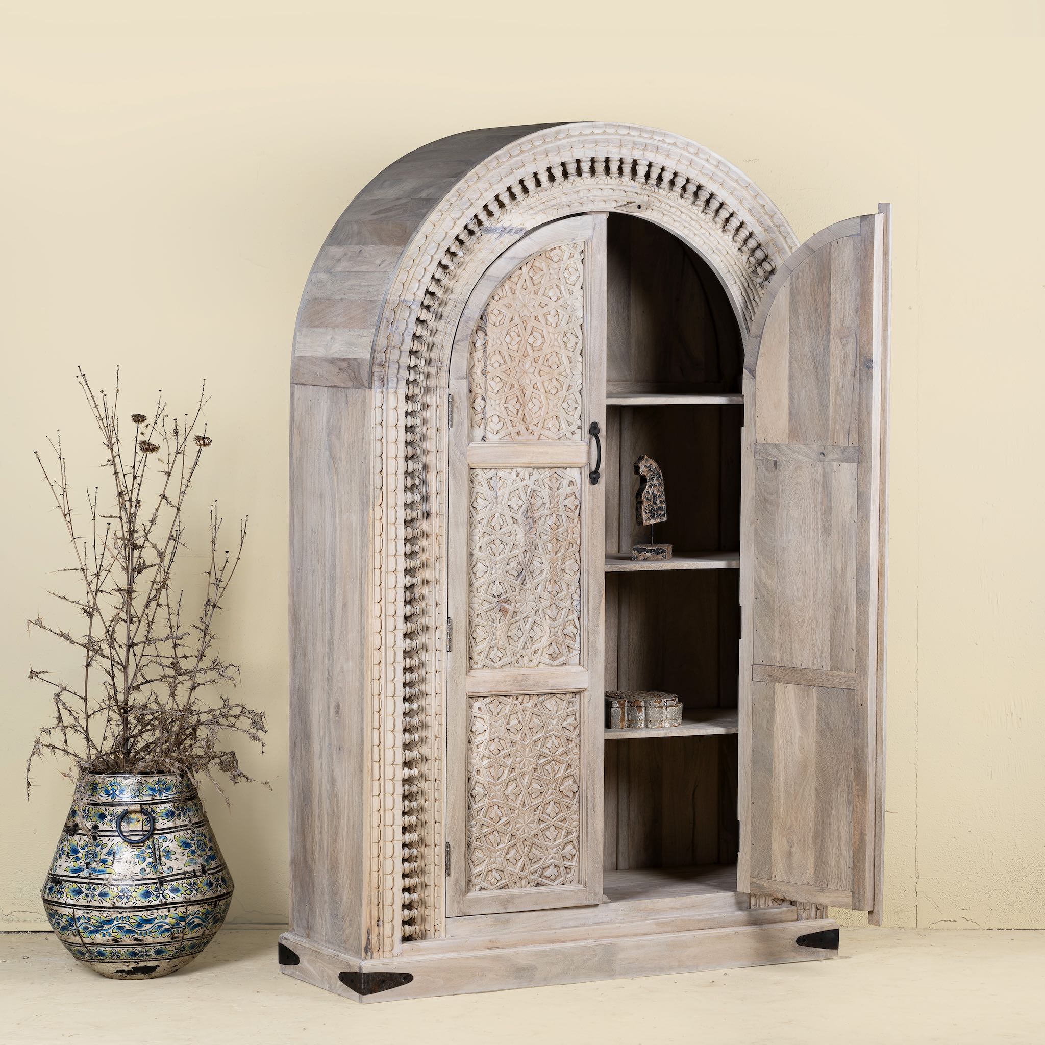
Side angle with open door of a hand-carved mango wood cabinet with an arched half-round top and ornate closed floral pattern doors, finished in a soft natural tone for timeless elegance.