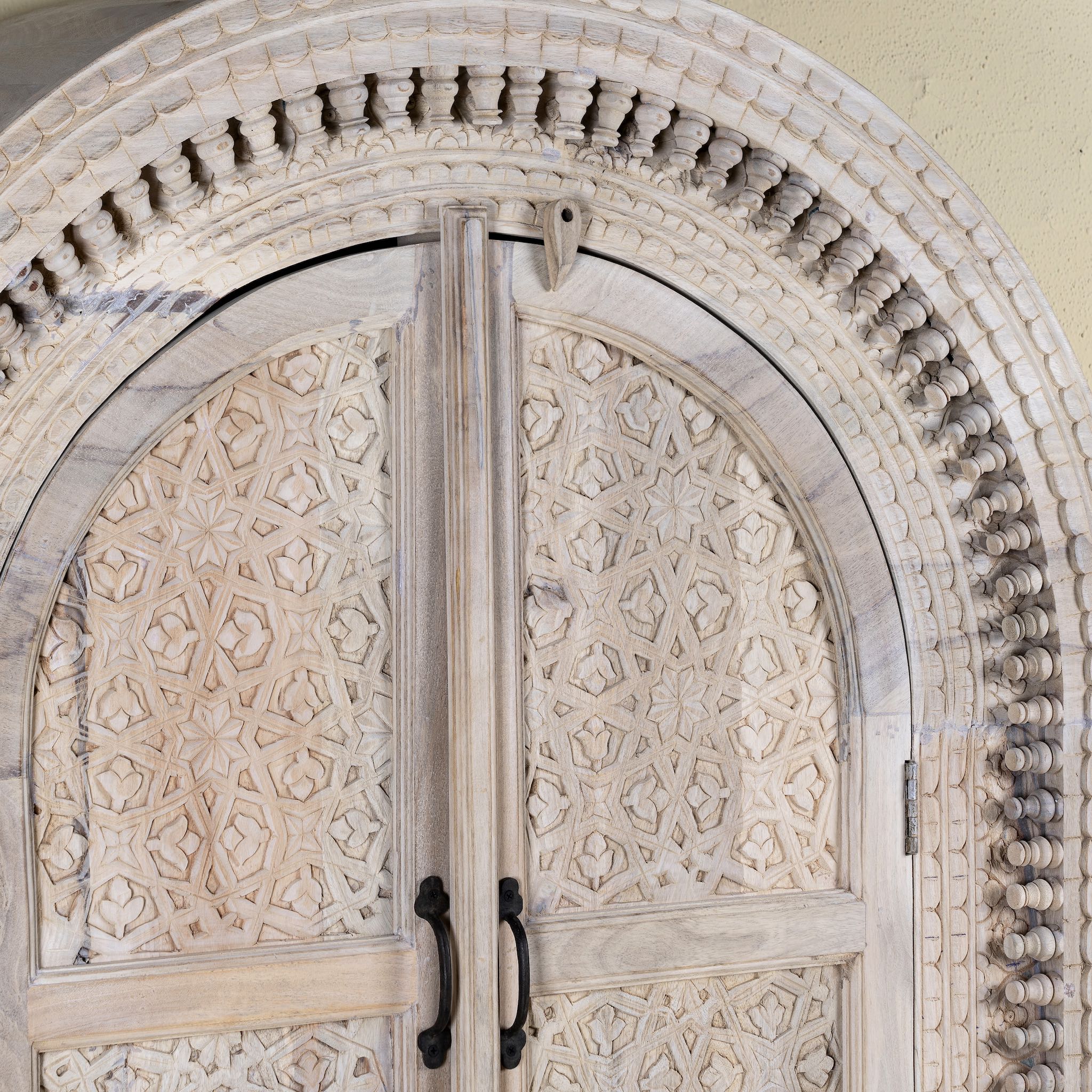 Close up picture of a hand-carved mango wood cabinet with an arched half-round top and ornate closed floral pattern doors, finished in a soft natural tone for timeless elegance.