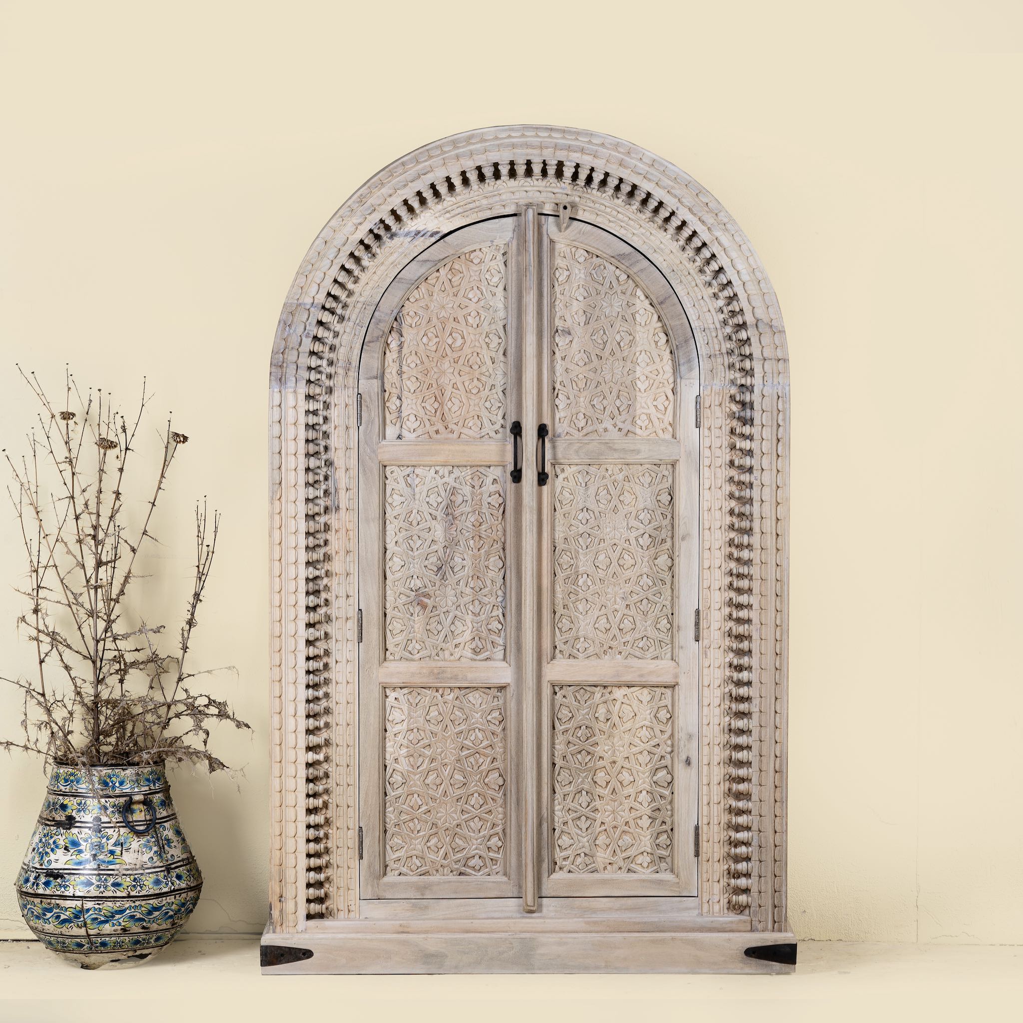 Hand-carved mango wood cabinet with an arched half-round top and ornate closed floral pattern doors, finished in a soft natural tone for timeless elegance.