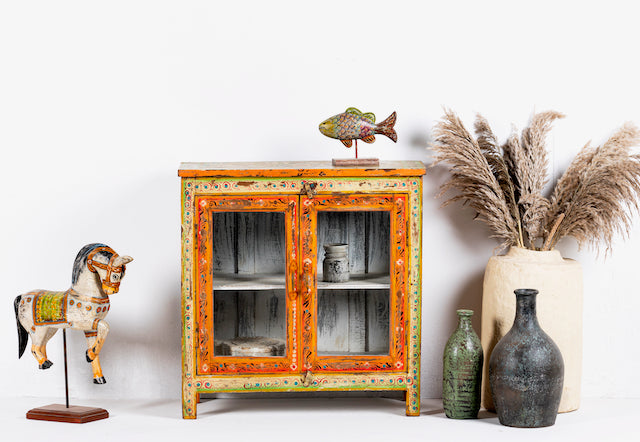 A hand-painted vintage showcase with floral and geometric patterns in orange, yellow, and green, made from refurbished old wood. The showcase has two glass panelled doors and is placed in a room with decorative items around it.