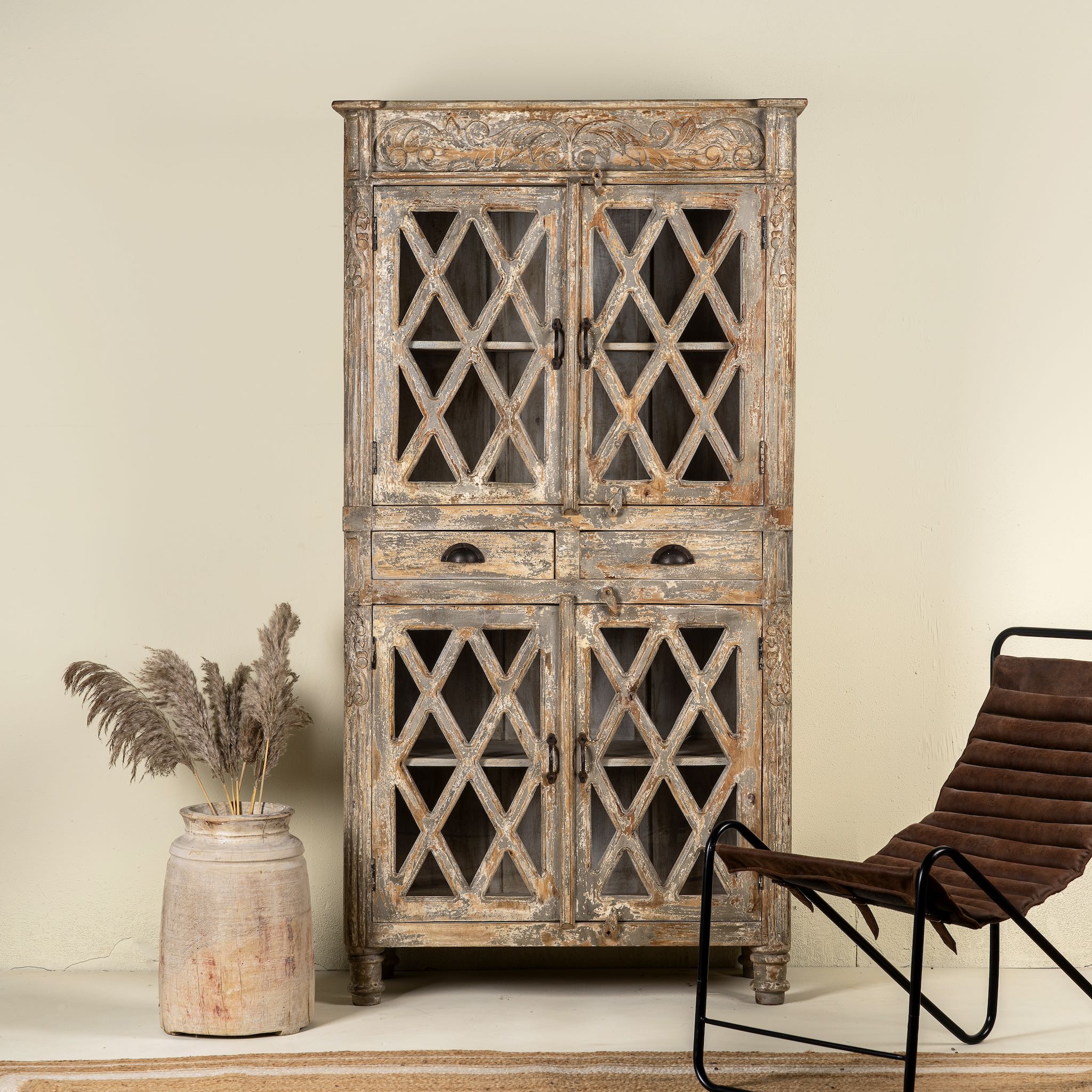 Handcrafted mango wood showcase cabinet with a rustic design, featuring glass-paneled doors and intricate wood carvings, placed in a room with a chair and decorative vase.