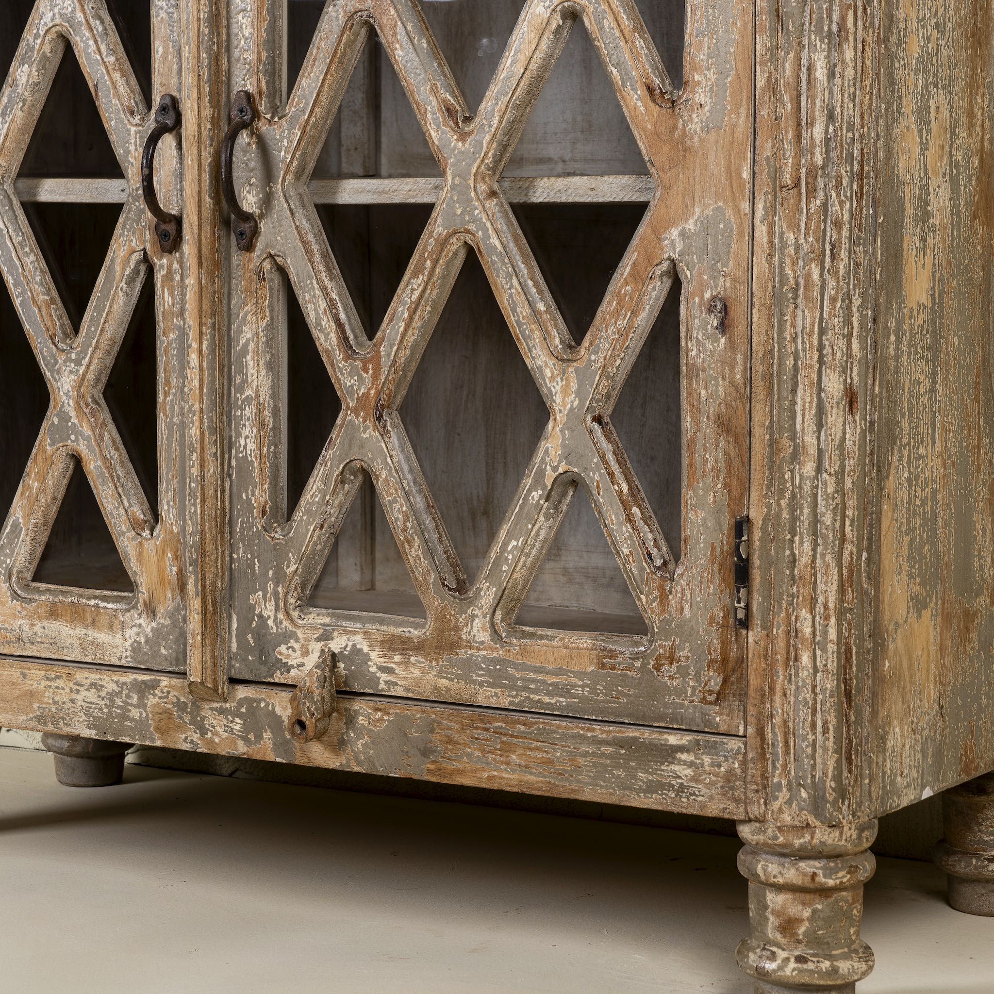 Close up image of the leg part of a handcrafted showcase cabinet made from reclaimed wood, featuring diamond-pattern glass pane doors, a crackled paint finish in earthy tones, two drawers, and vintage-inspired detailing.