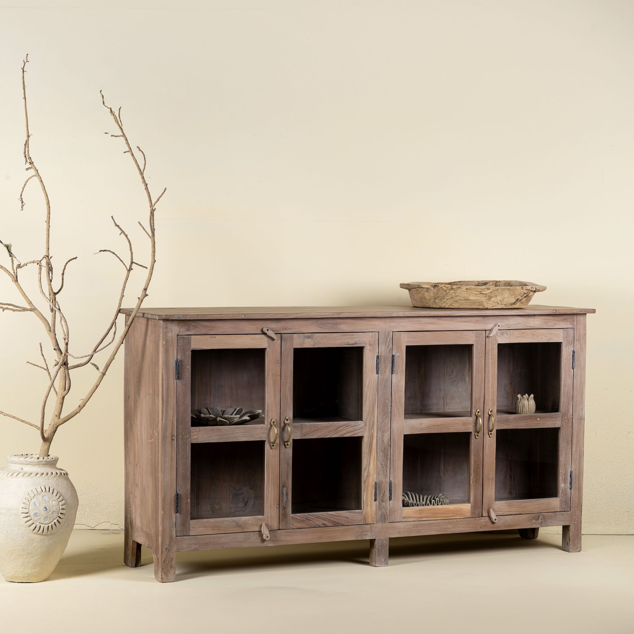 Side angle image of a rustic showcase cabinet made from recycled teak wood, featuring four glass-paneled doors with brass handles and a lightly weathered finish, offering a warm, natural look ideal for displaying decor or tableware.
