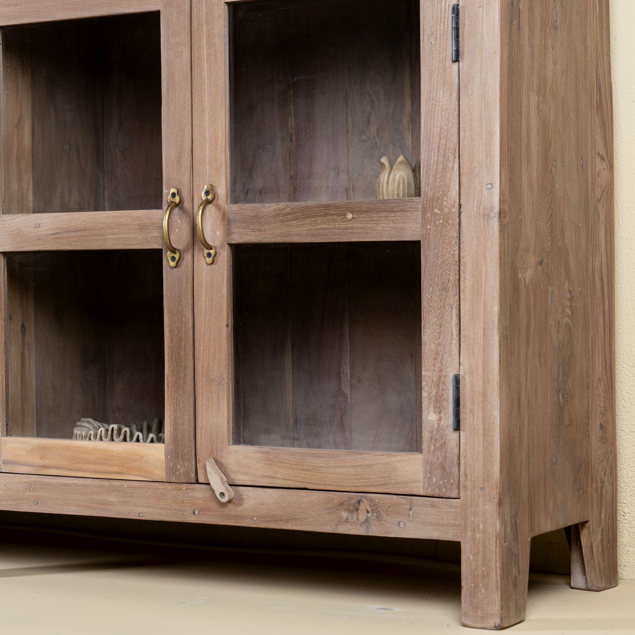 Close up image of the leg of a rustic showcase cabinet made from recycled teak wood, featuring four glass-paneled doors with brass handles and a lightly weathered finish, offering a warm, natural look ideal for displaying decor or tableware.
