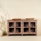 Front  view image of a rustic showcase cabinet made from recycled teak wood, featuring four glass-paneled doors with brass handles and a lightly weathered finish, offering a warm, natural look ideal for displaying decor or tableware.
