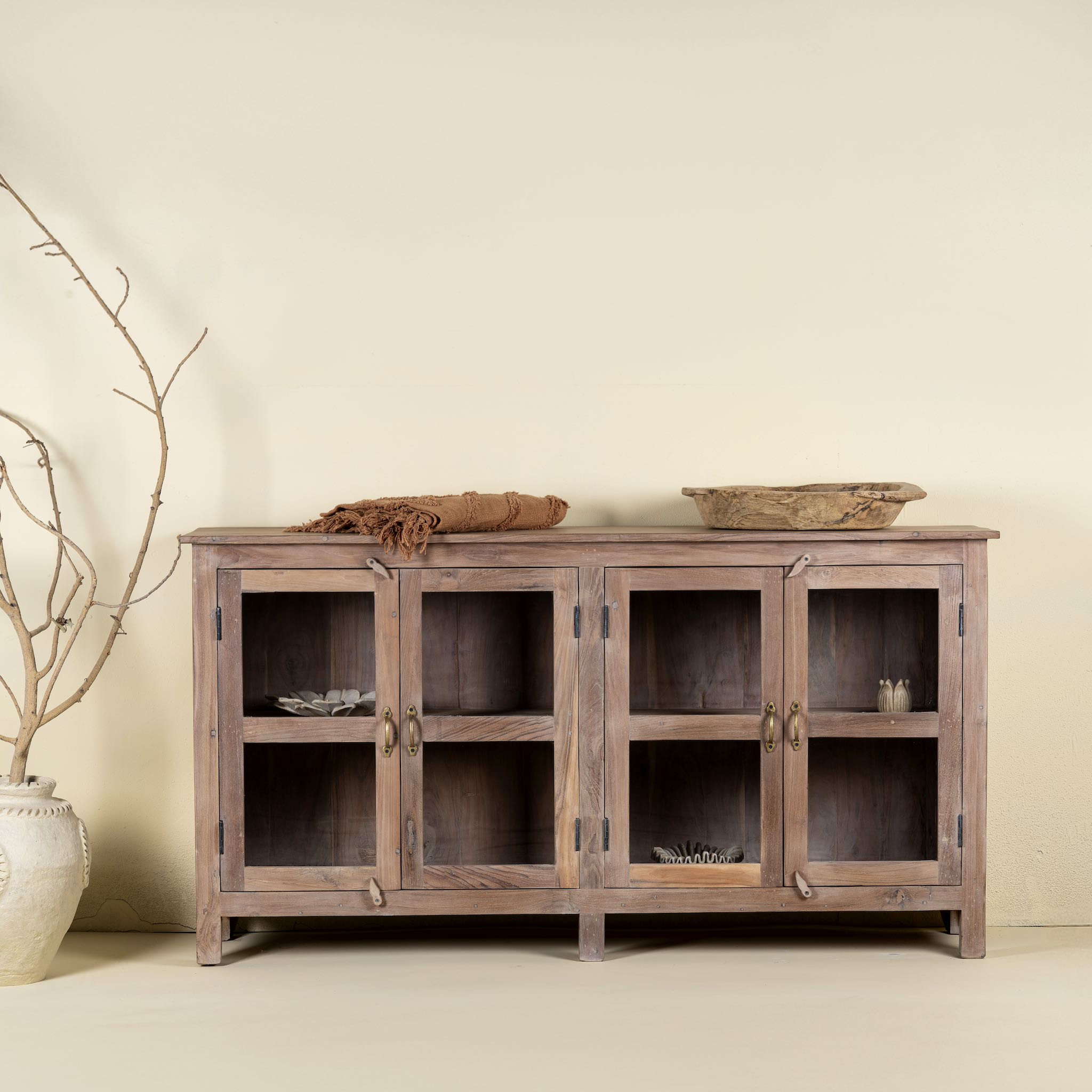 Front  view image of a rustic showcase cabinet made from recycled teak wood, featuring four glass-paneled doors with brass handles and a lightly weathered finish, offering a warm, natural look ideal for displaying decor or tableware.
