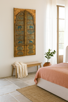 Lifestyle scene of a hand-painted wooden wall panel made from an old window frame, featuring intricate blue floral patterns along with stylised animal motifs such as elephants and camels, set against a distressed golden-brown background with an arched top frame.
