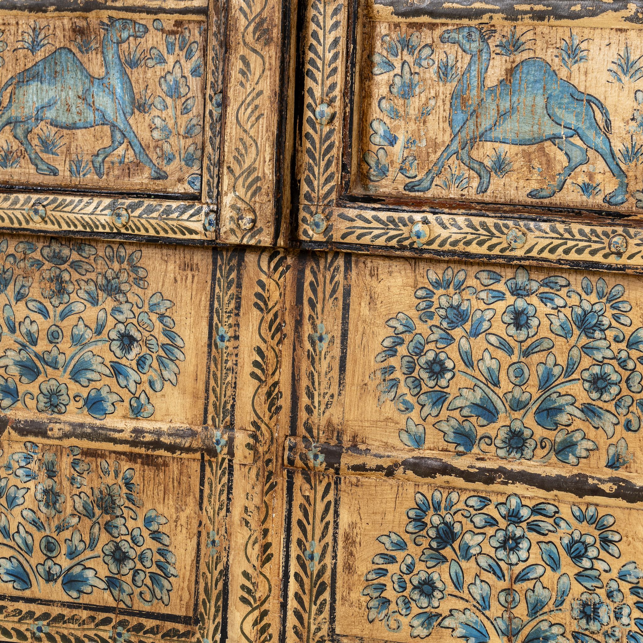 Close up image of a hand-painted wooden wall panel made from an old window frame, featuring intricate blue floral patterns along with stylised animal motifs such as elephants and camels, set against a distressed golden-brown background with an arched top frame.
